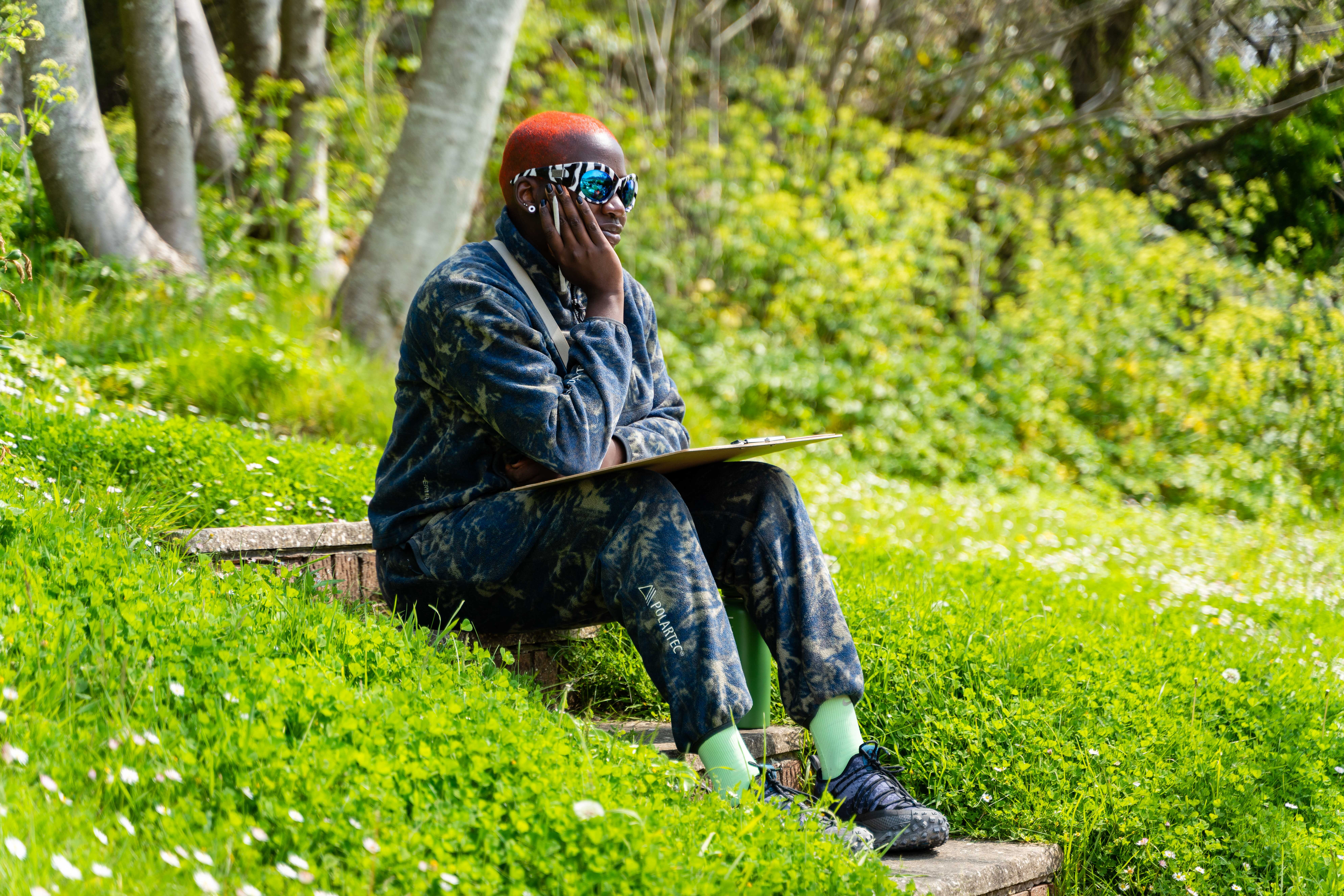 Person in dark clothing and red cap sits on wooden bench reading, surrounded by bright green grass and trees in background.