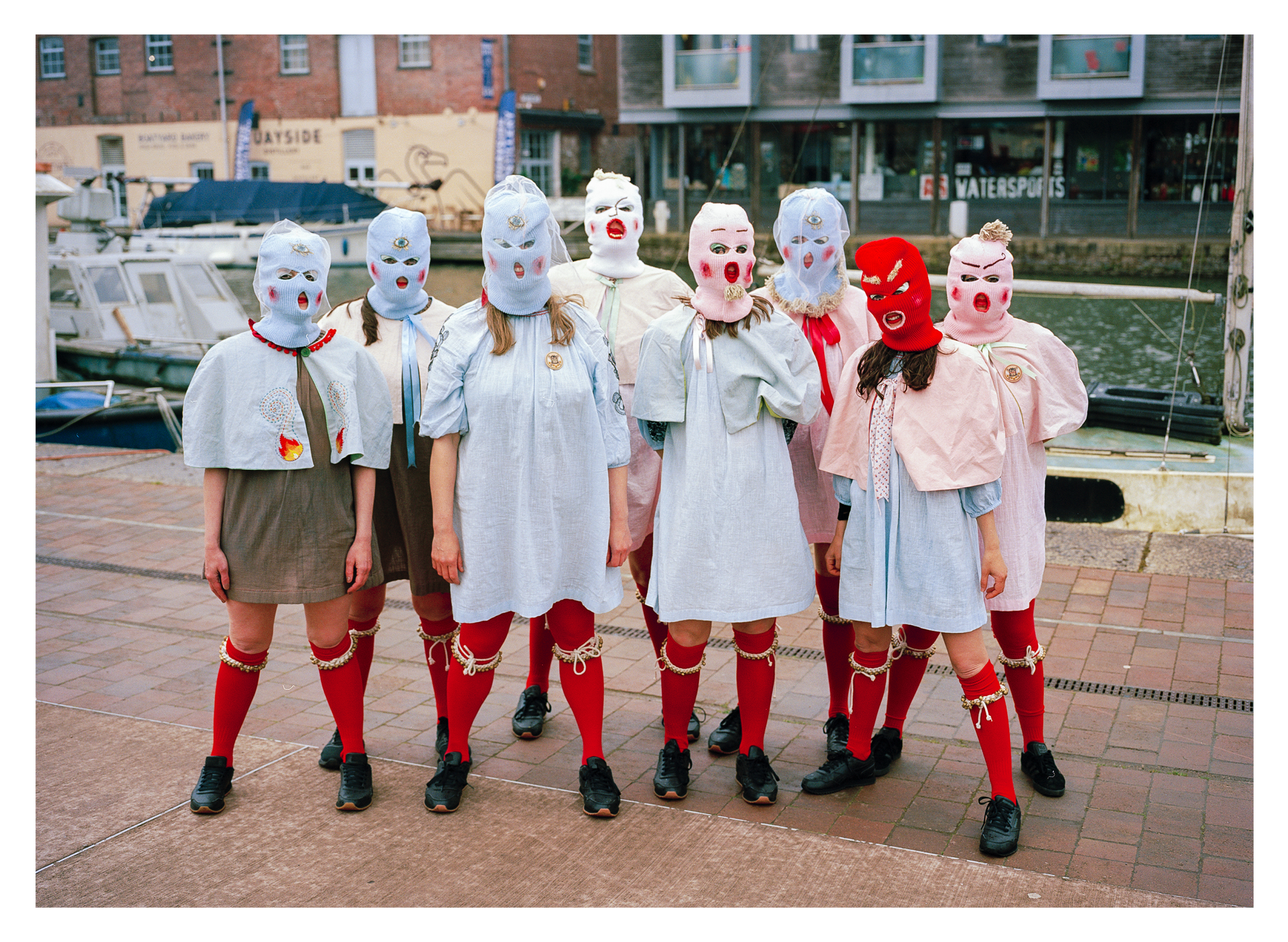 Seven people wearing white masks and red socks standing on brick pavement in front of shops and parked cars.