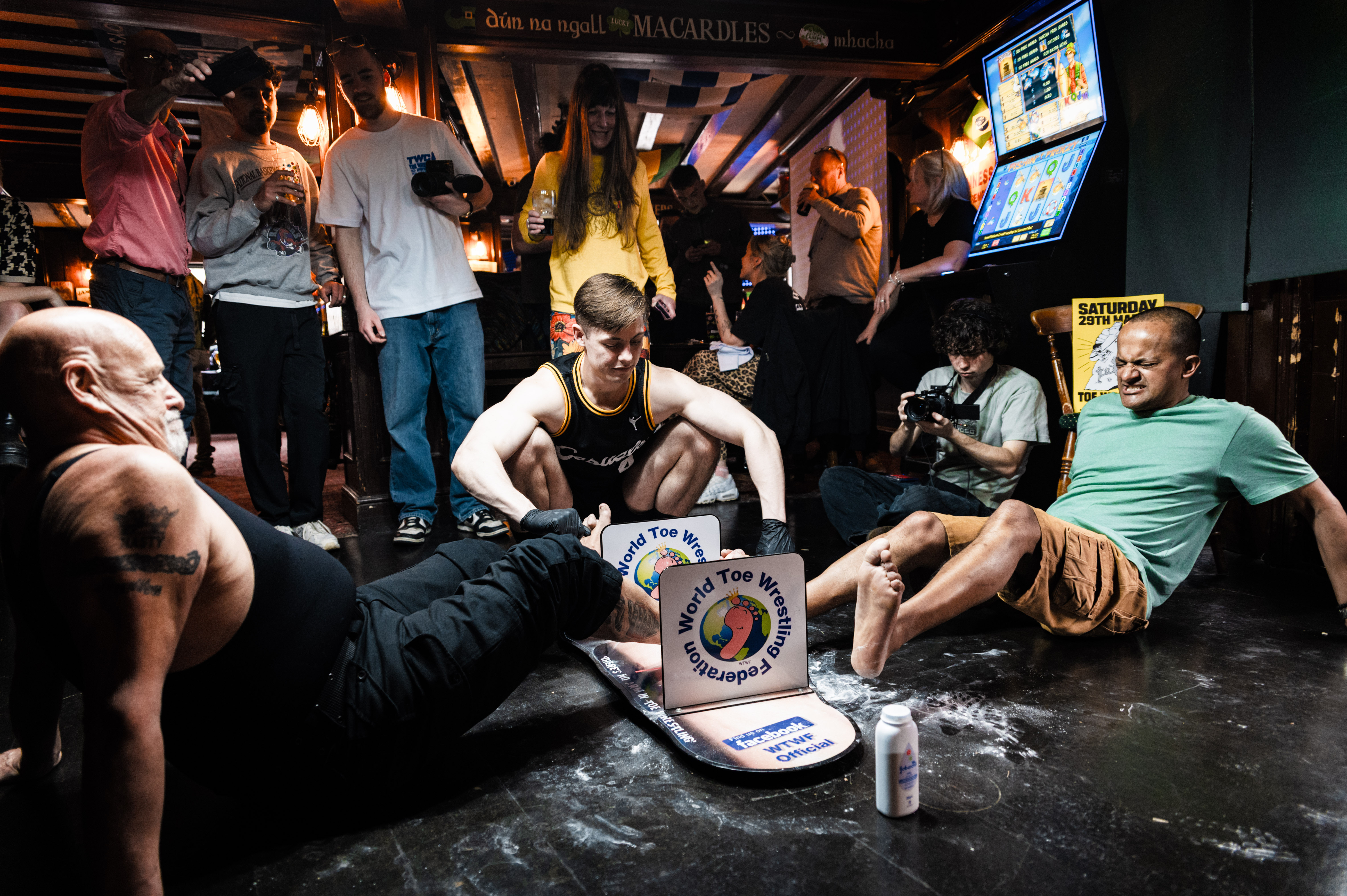 Crowded pub with people sitting on the floor, some holding drinks. Assorted items on the floor including a sign and a bottle.