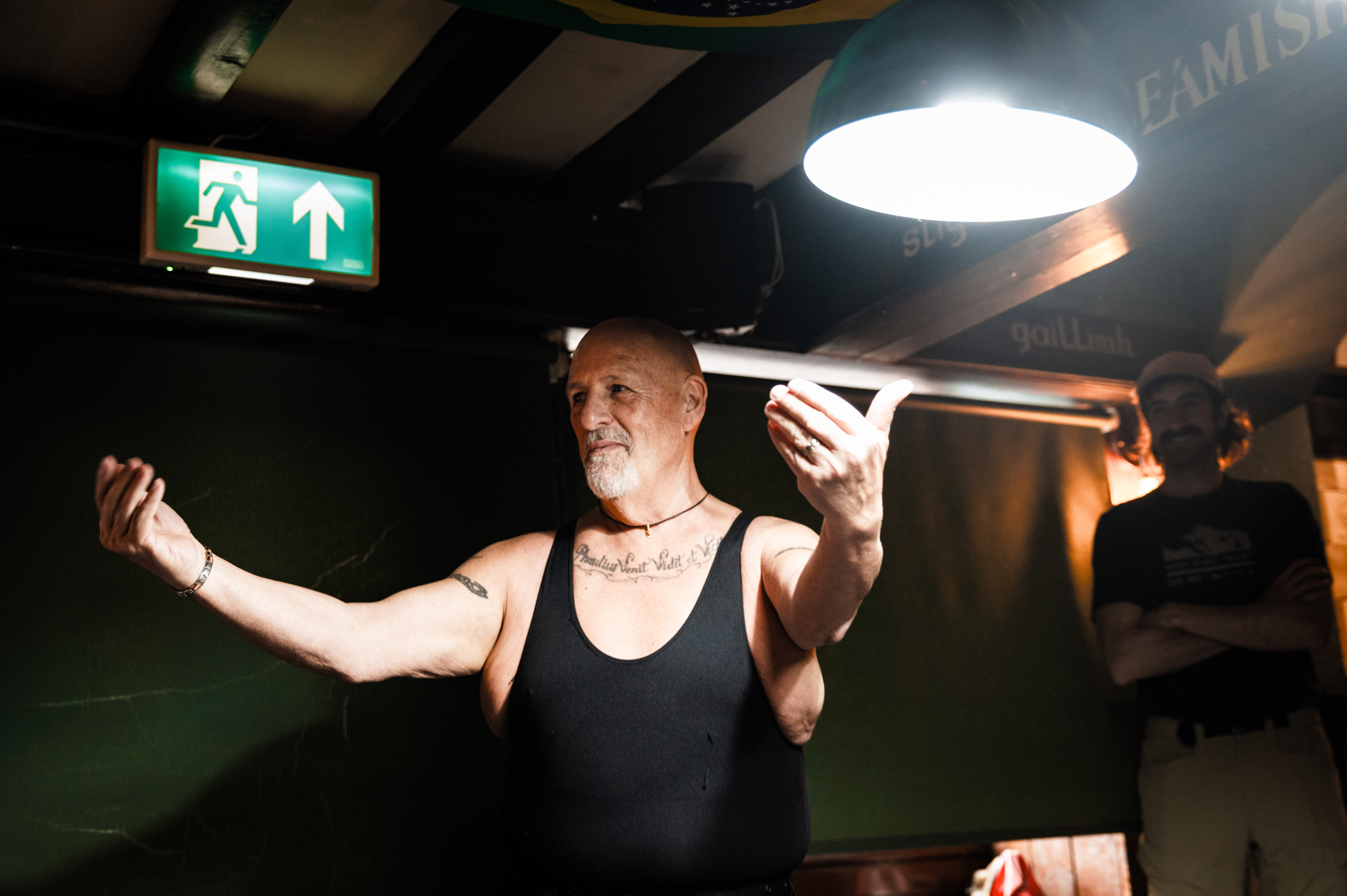 A man in a black singlet gesturing with his arms raised in a dimly lit room with an exit sign visible.