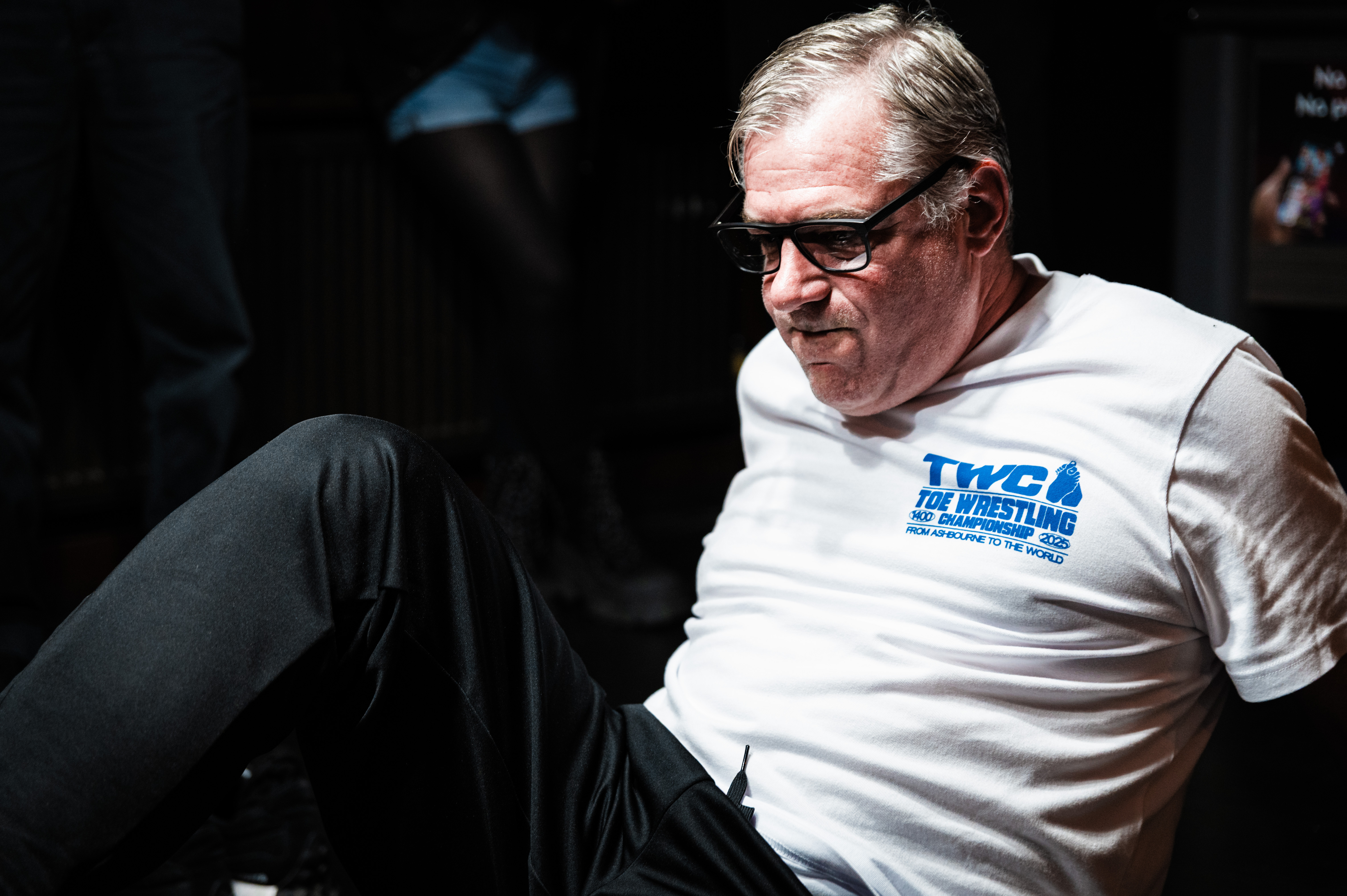 An older man with grey hair wearing a white T-shirt with TW2L Wrestling printed on it, sitting in a dark setting.