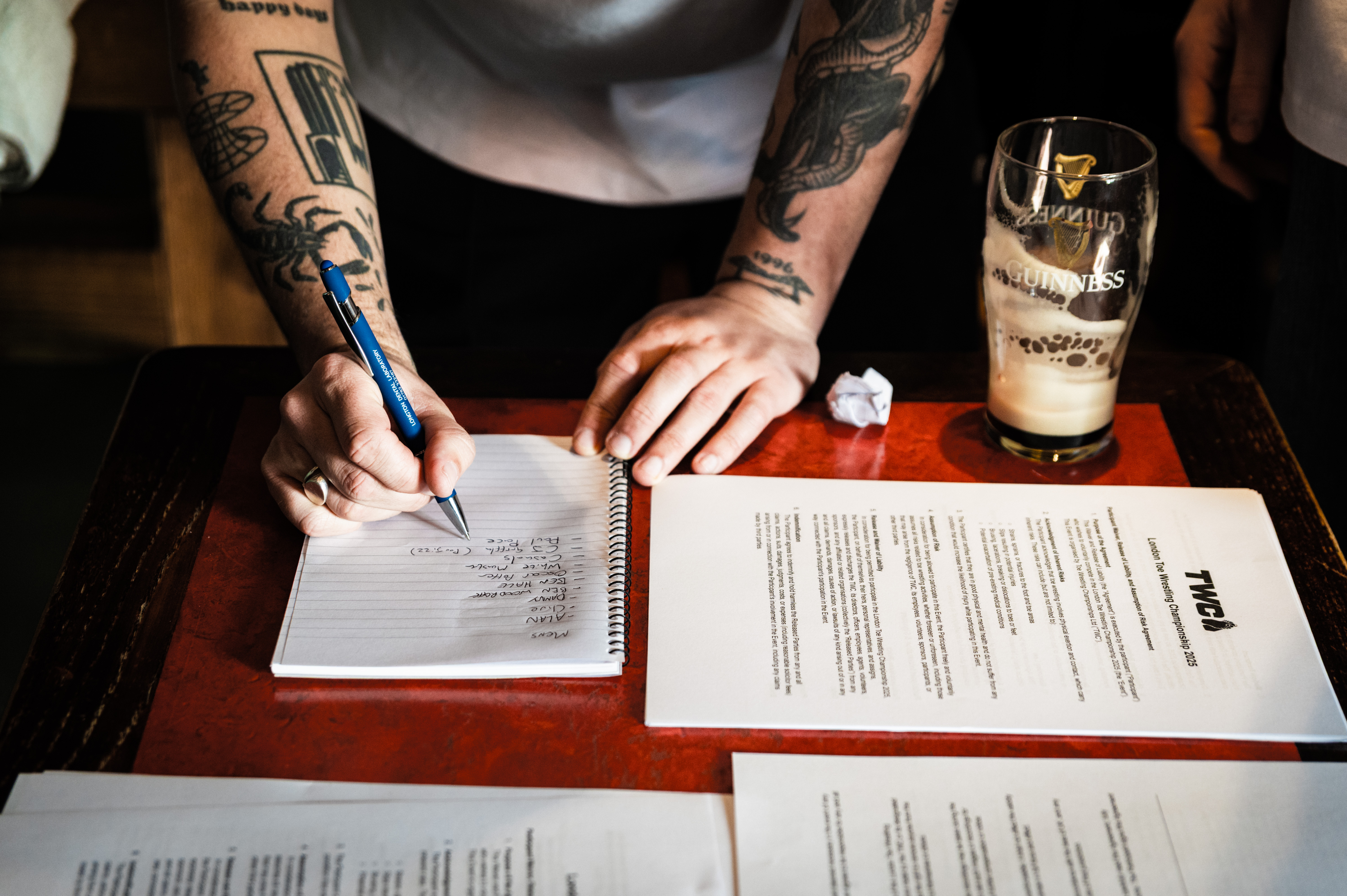 Tattooed hands holding pen, writing on document with pint of beer on table.