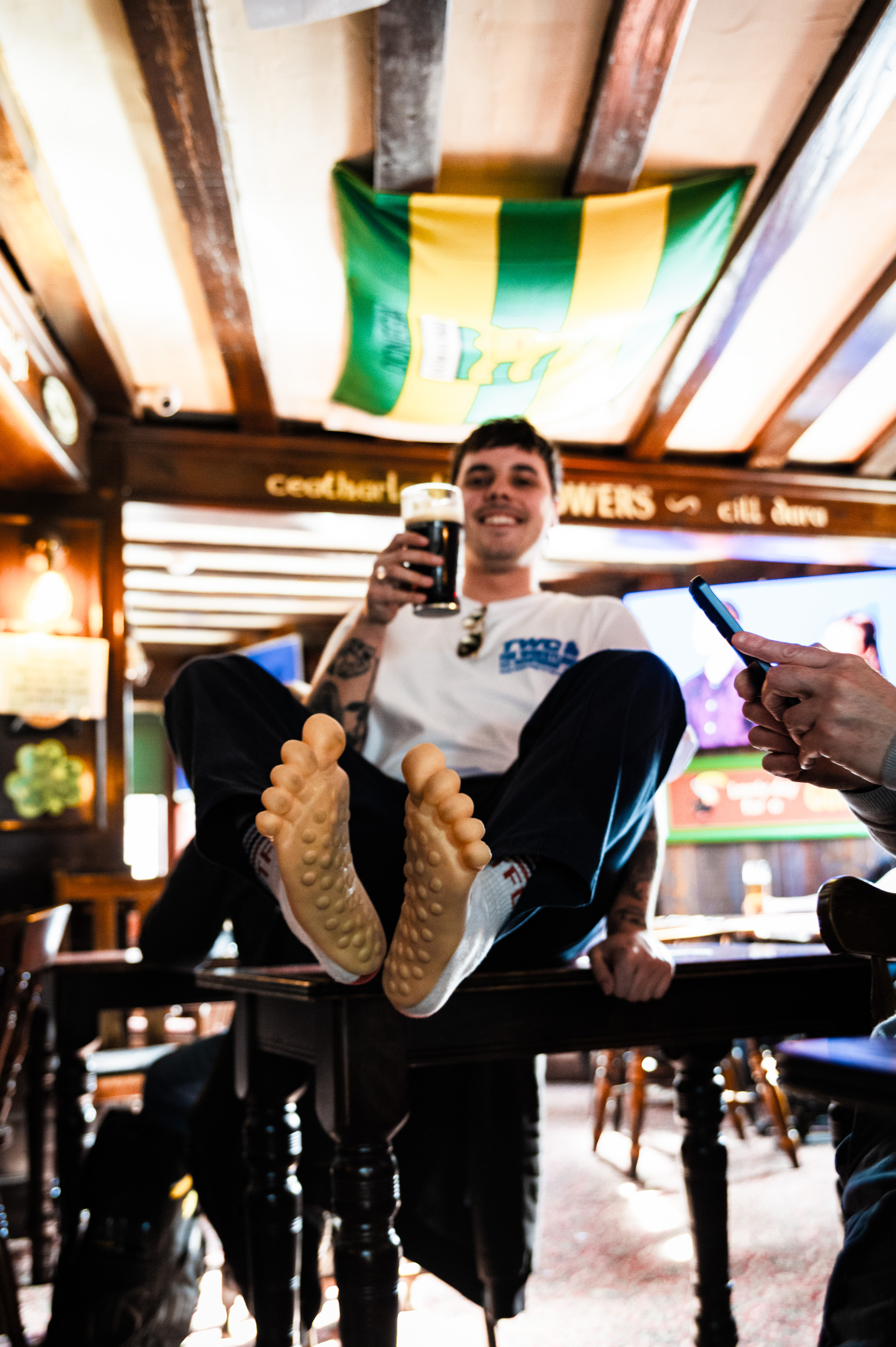 A man wearing a white T-shirt and black trousers sitting in a bar, holding a drink and showing his shoes.