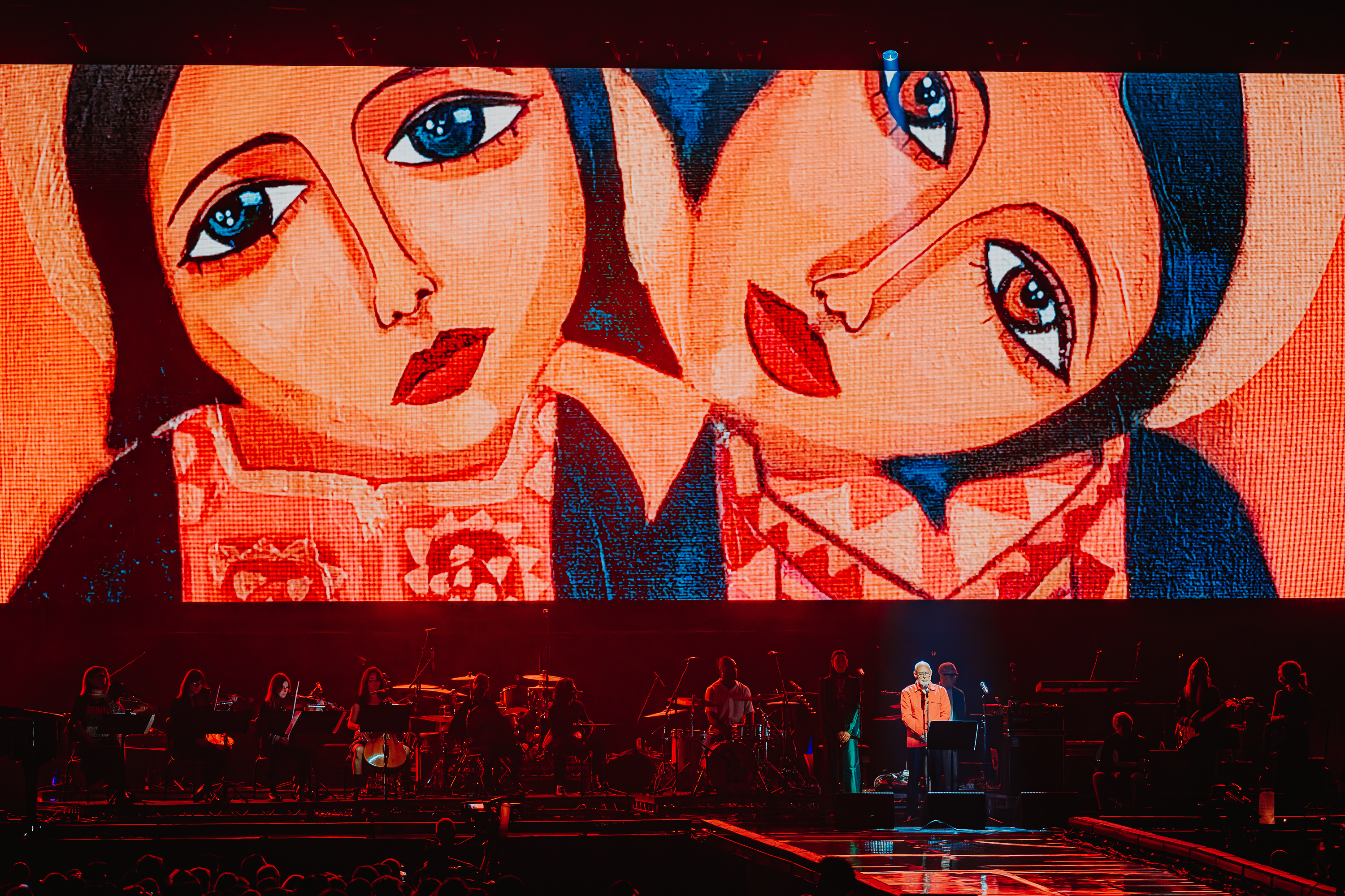 Large screen displays stylised illustration of two figures with orange skin, blue eyes, red lips, and dark hair. Performer on stage below.