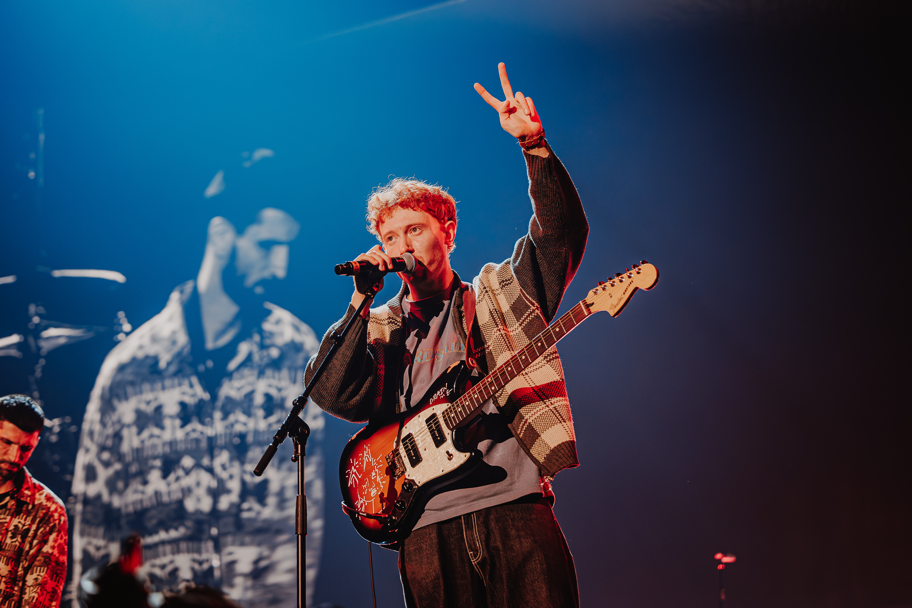 Musician with blonde hair holding electric guitar and raising left hand whilst singing into microphone on stage with blue lighting.