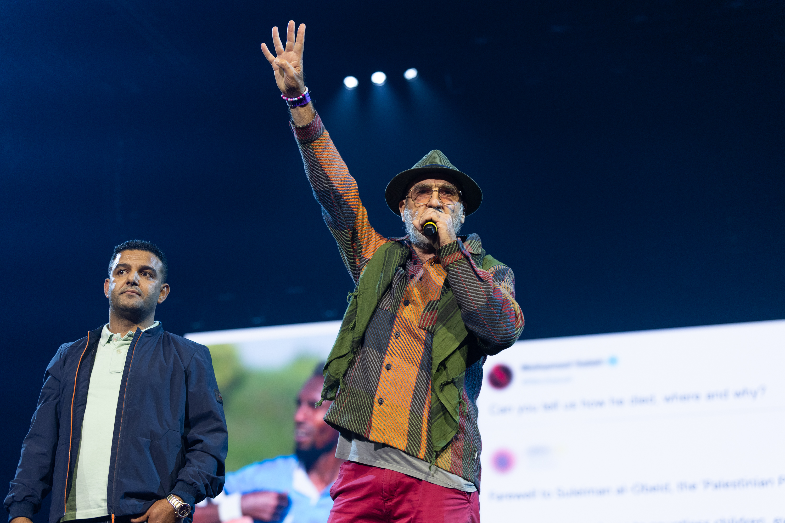 Two men on stage with blue lighting, one in dark suit, other in hat and colourful jacket holding microphone with raised arm.