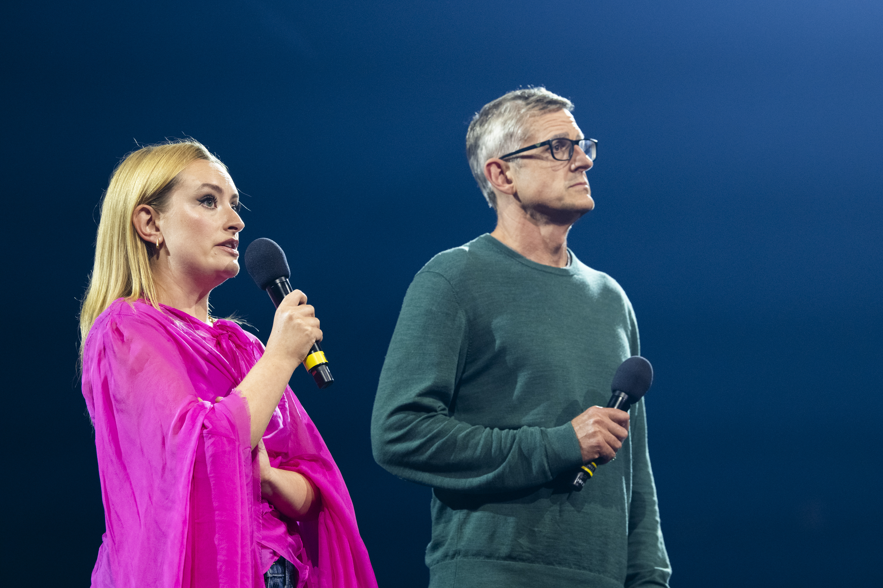 Woman in bright pink top holds microphone beside man in green jumper and glasses against blue backdrop.