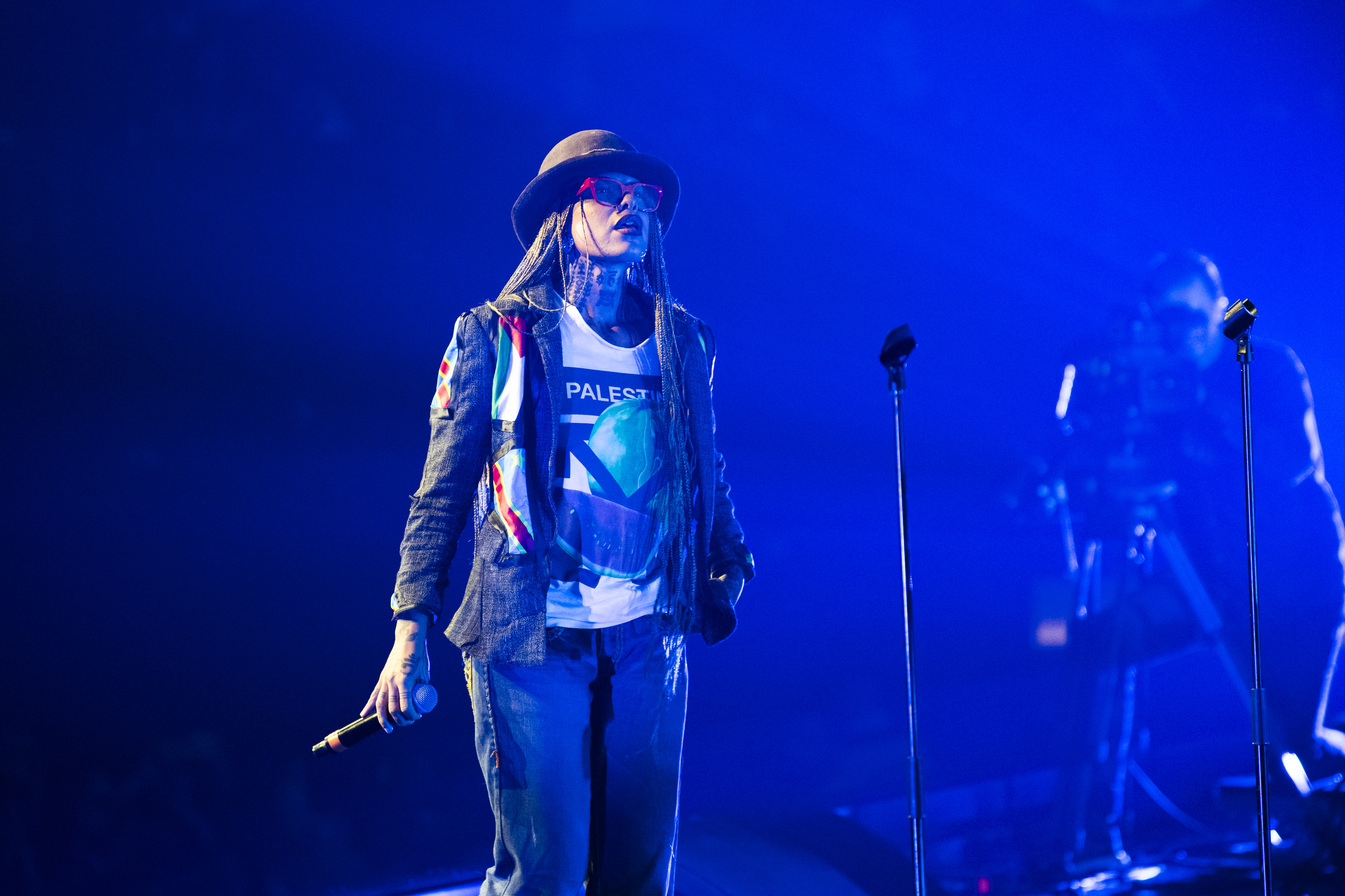 Performer in denim jacket and graphic t-shirt on stage with microphone stands, bathed in deep blue stage lighting.