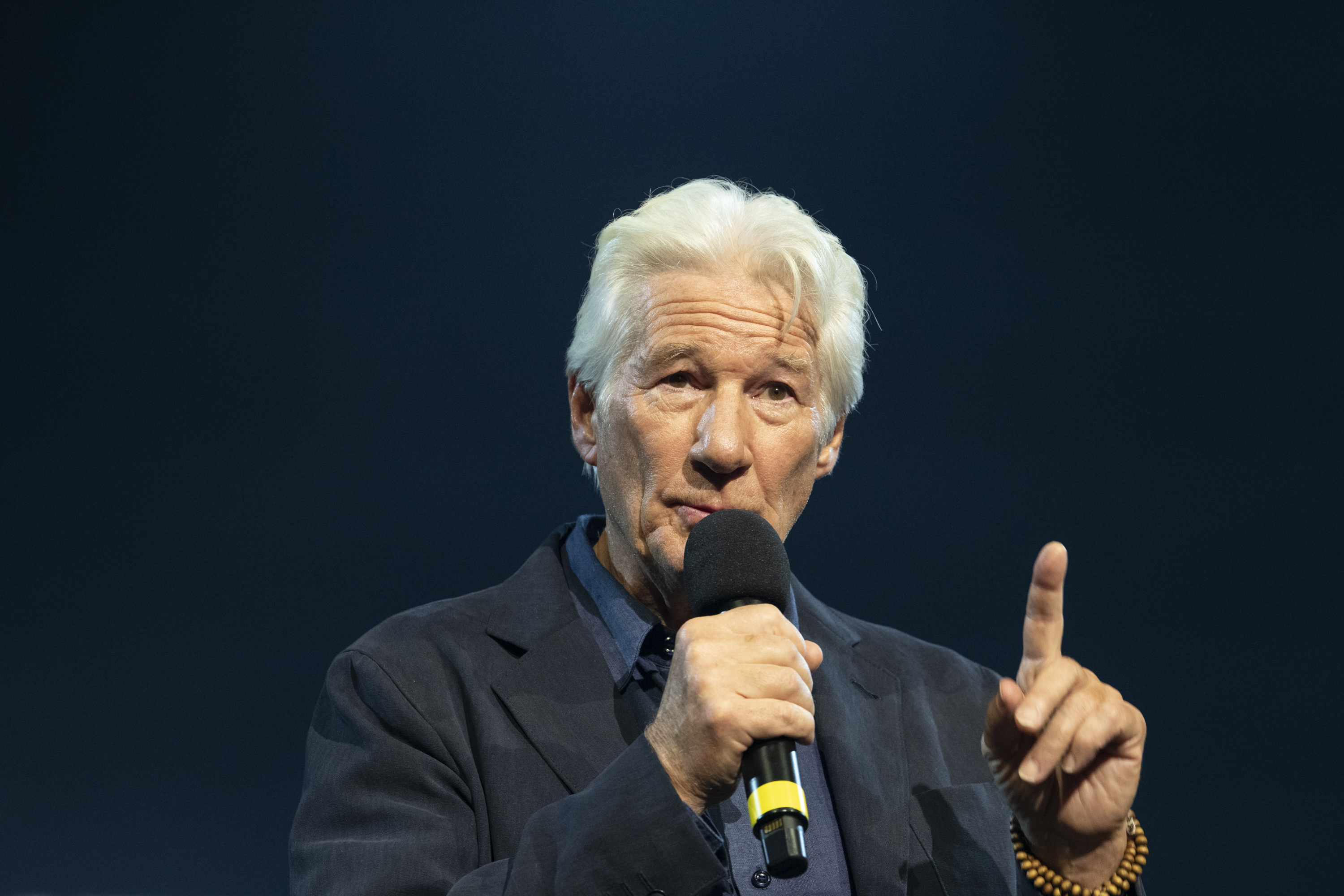 Elderly man with grey hair speaking into black microphone, pointing finger upward, wearing dark suit against blue background.