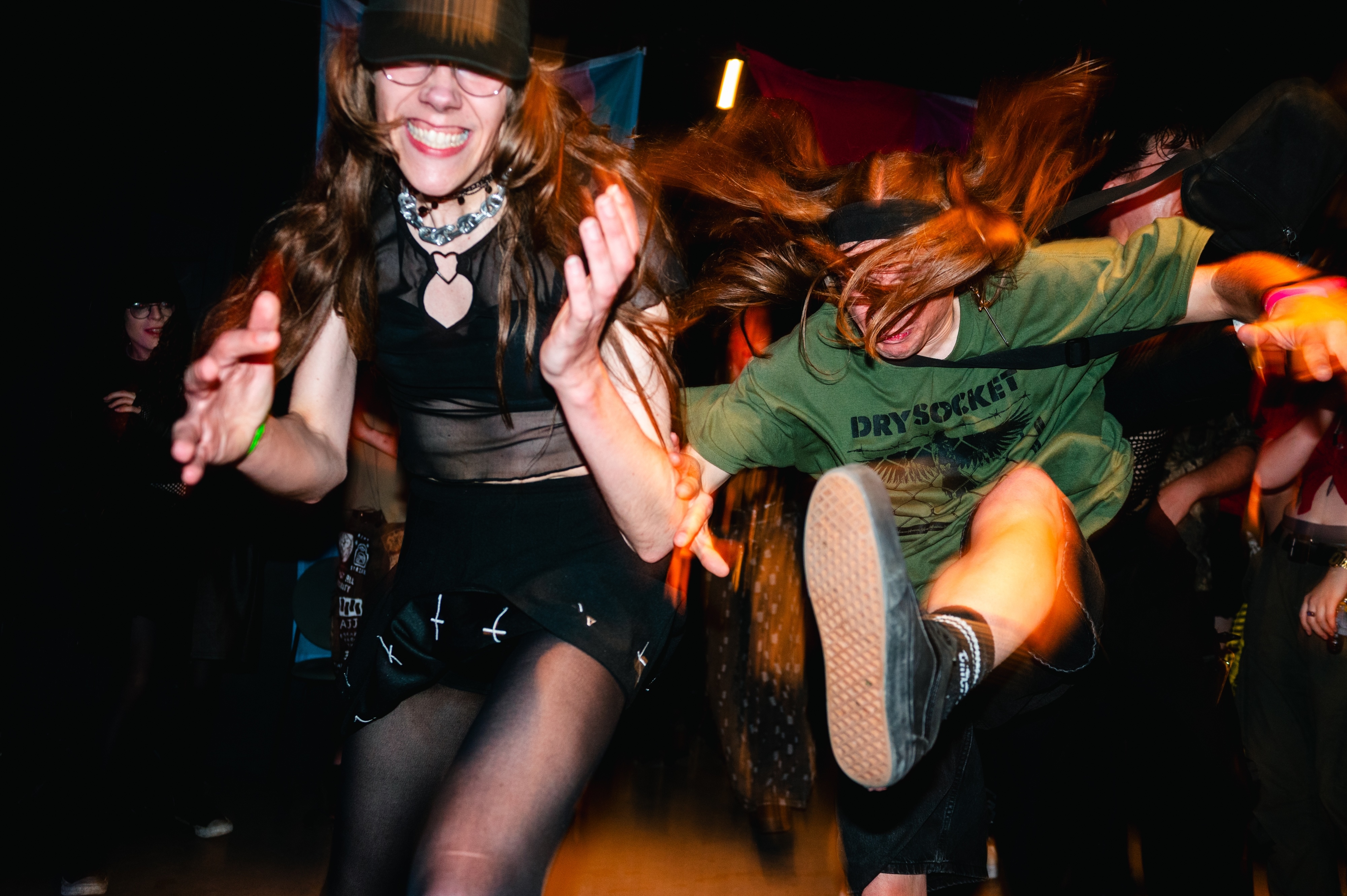 Woman in black top and fishnet tights dancing with arms raised, person in green shirt jumping behind her, crowded nightclub scene