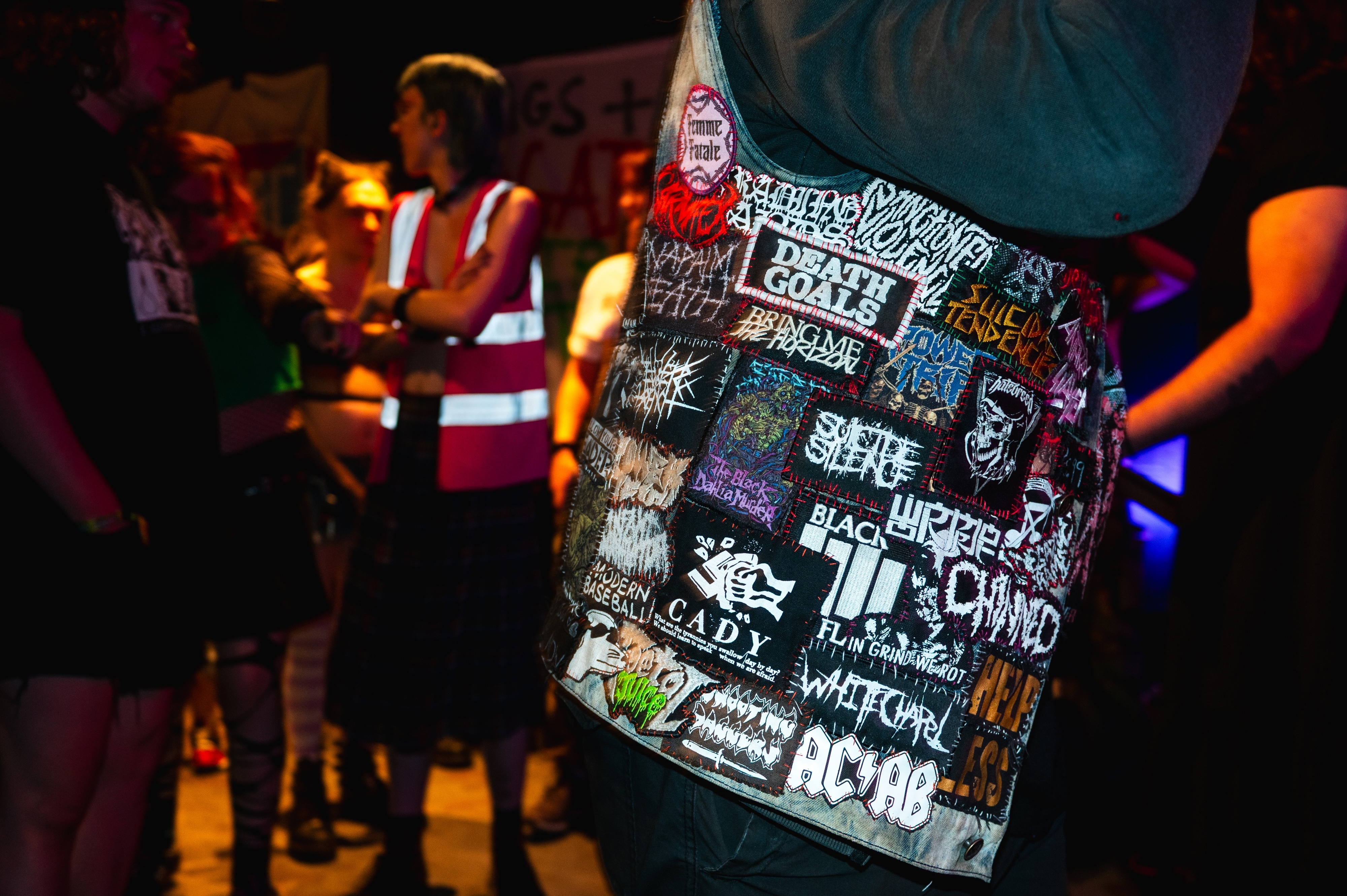 Person wearing black jacket covered in band patches and logos, with blurred figures in background under coloured lighting.