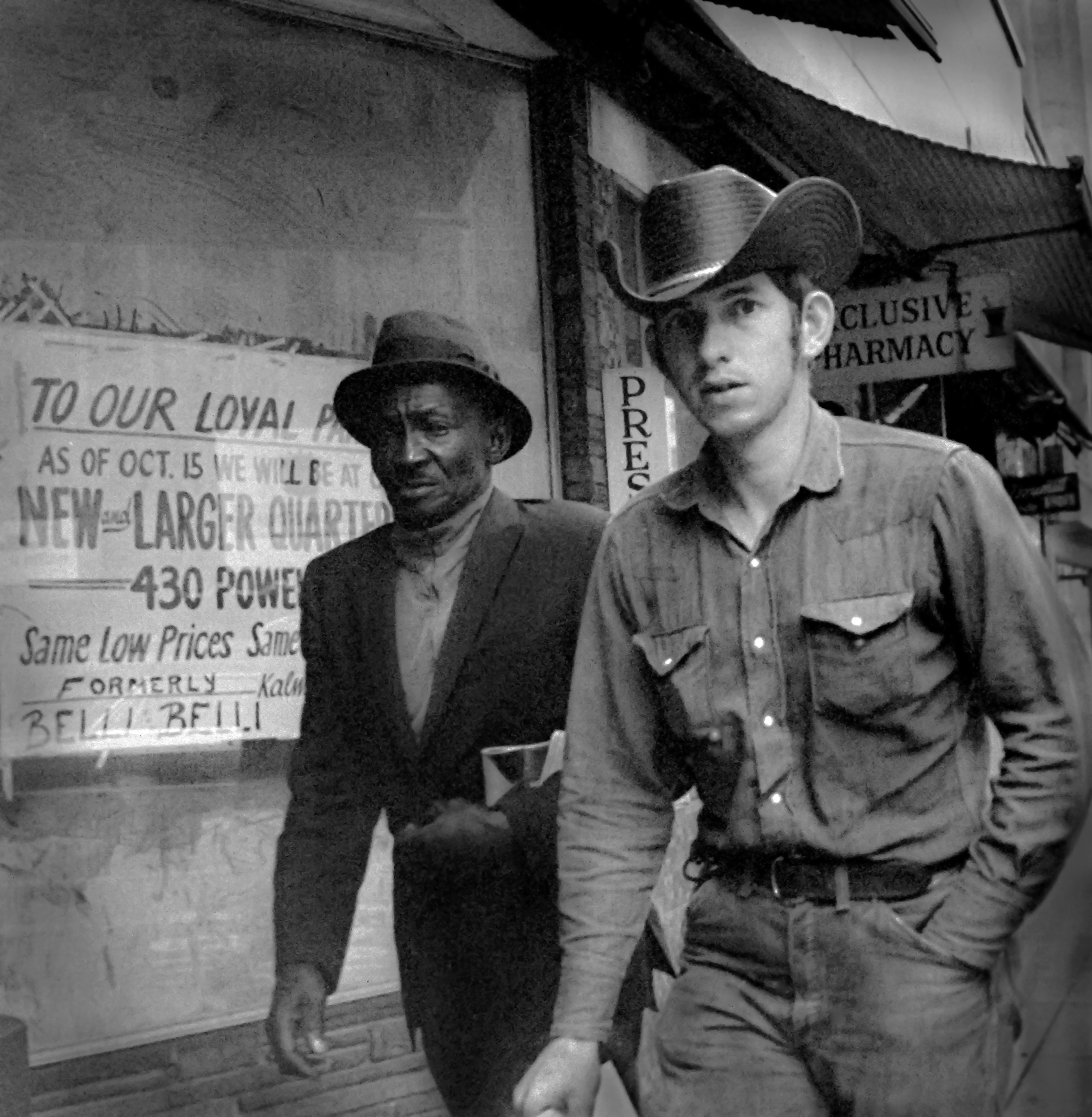 Two men, one older and wearing a hat, the other younger and in casual clothing, standing in front of a storefront with signage.