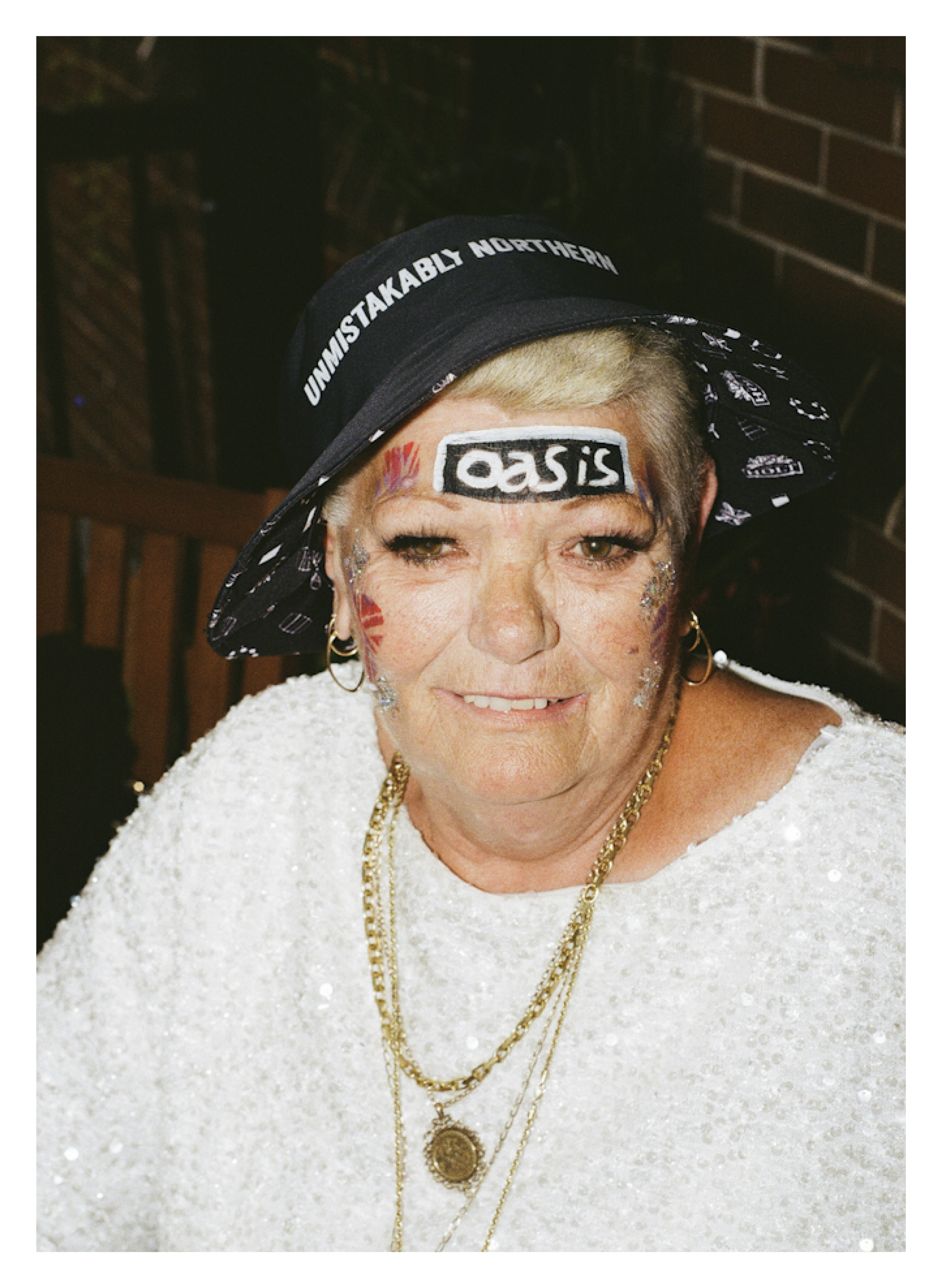 Person wearing black headband with white "OASIS" logo sticker on forehead, gold chains, and white top against brick wall background.