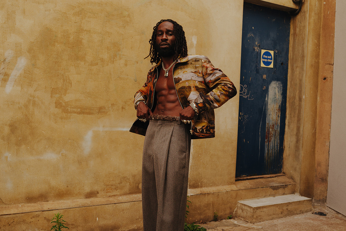 Man with dreadlocks wearing printed jacket and grey trousers stands against cream wall beside blue door with white paint marks.