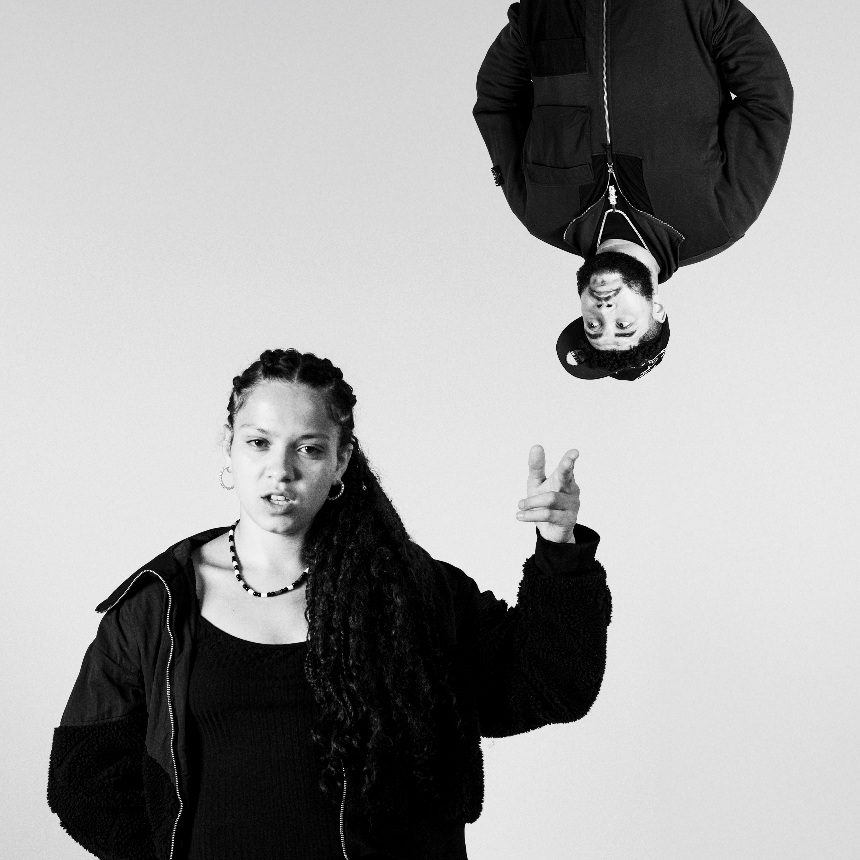 Black and white image of woman in dark jacket making peace sign, with upside-down person in black jacket above her.