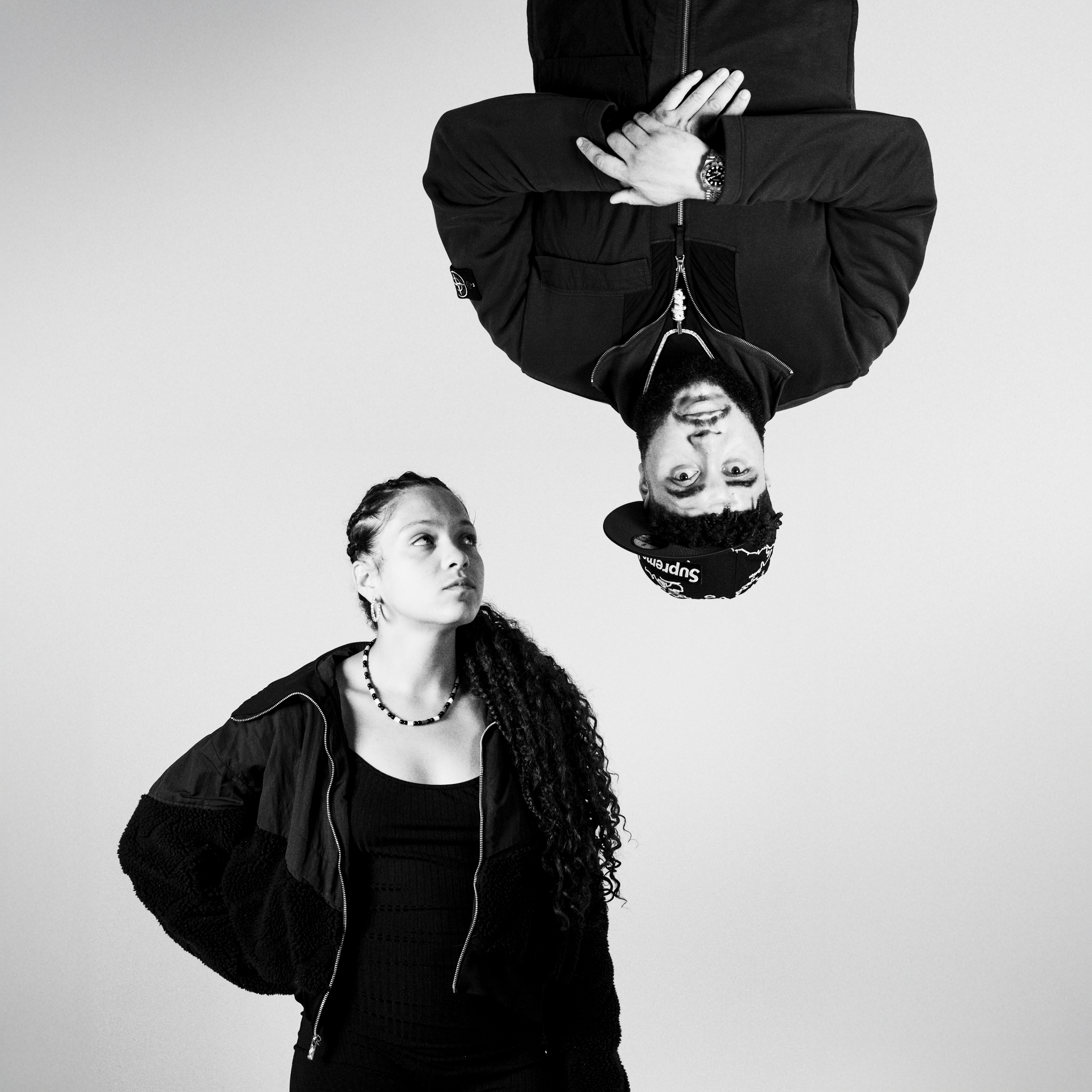 Black and white image of woman in dark jacket looking up at upside-down man in hoodie with hands behind head against white background.