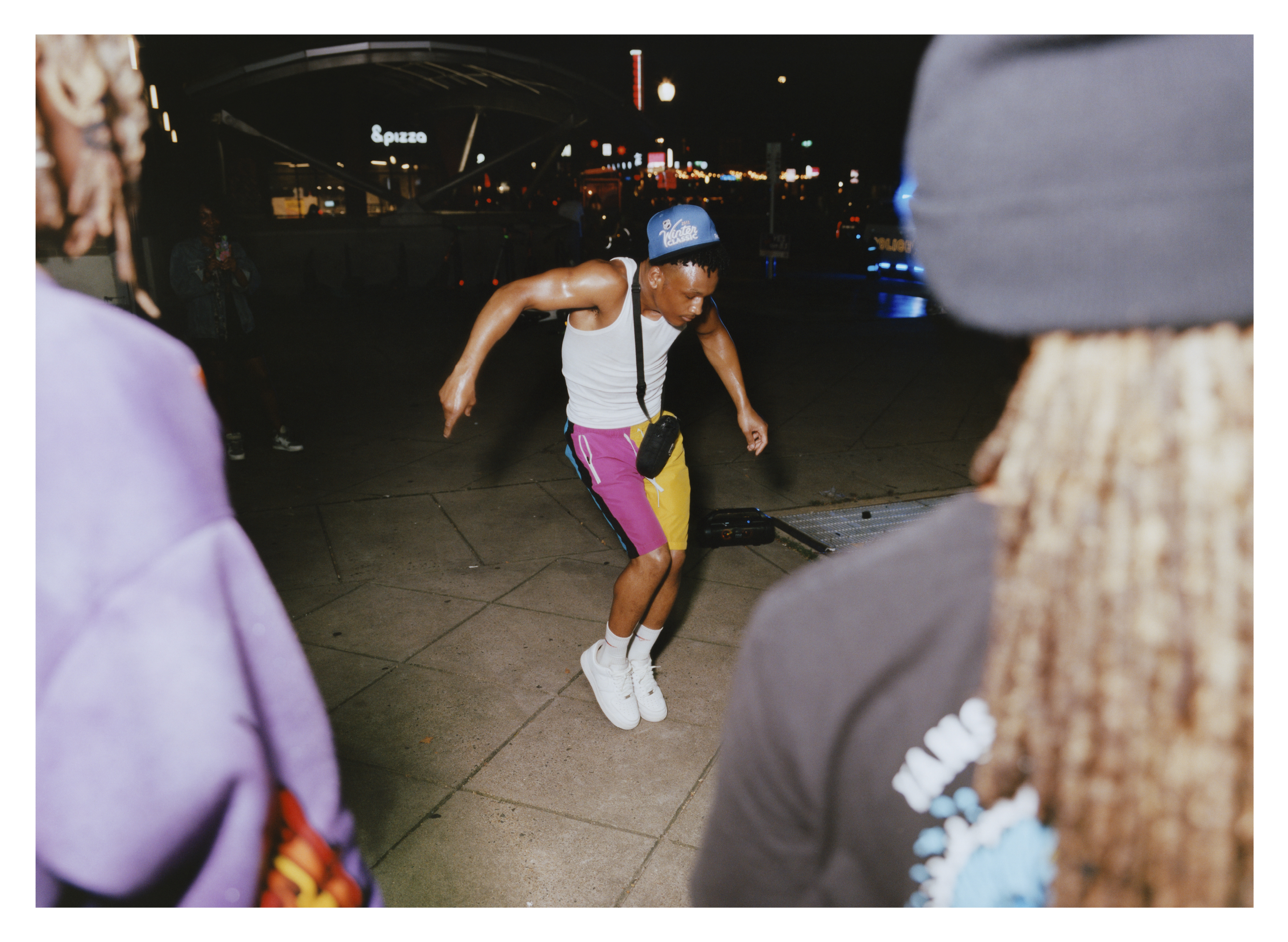 Person roller skating in white vest, pink and yellow shorts, and white skates on tiled floor at night with others nearby.