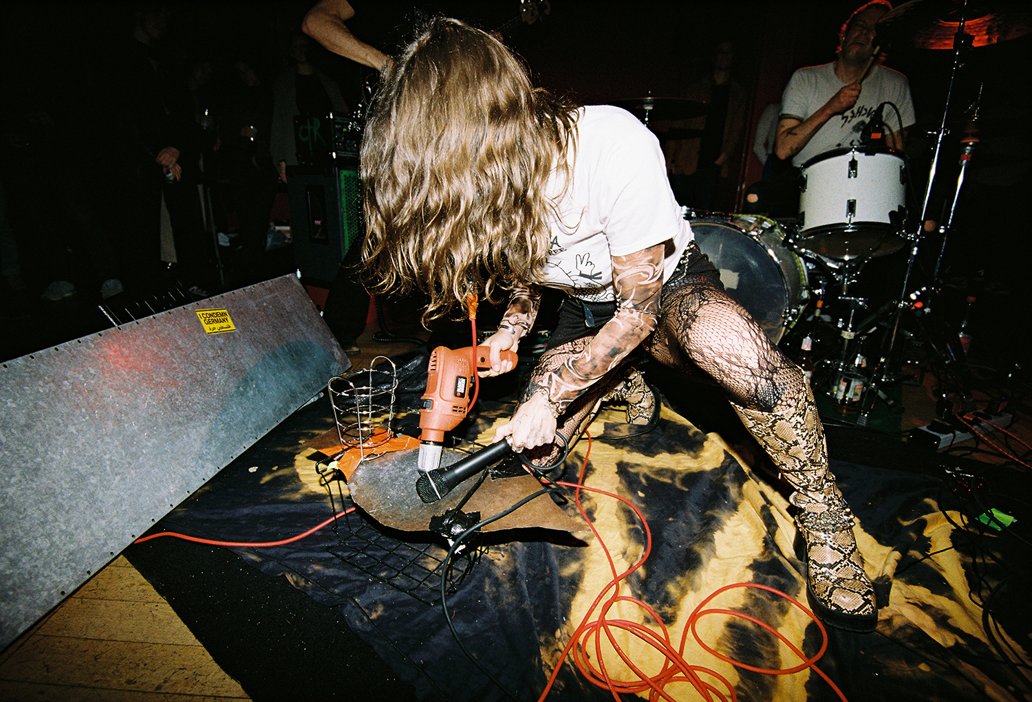 A person playing a guitar on stage, with long blonde curly hair and wearing a patterned outfit.