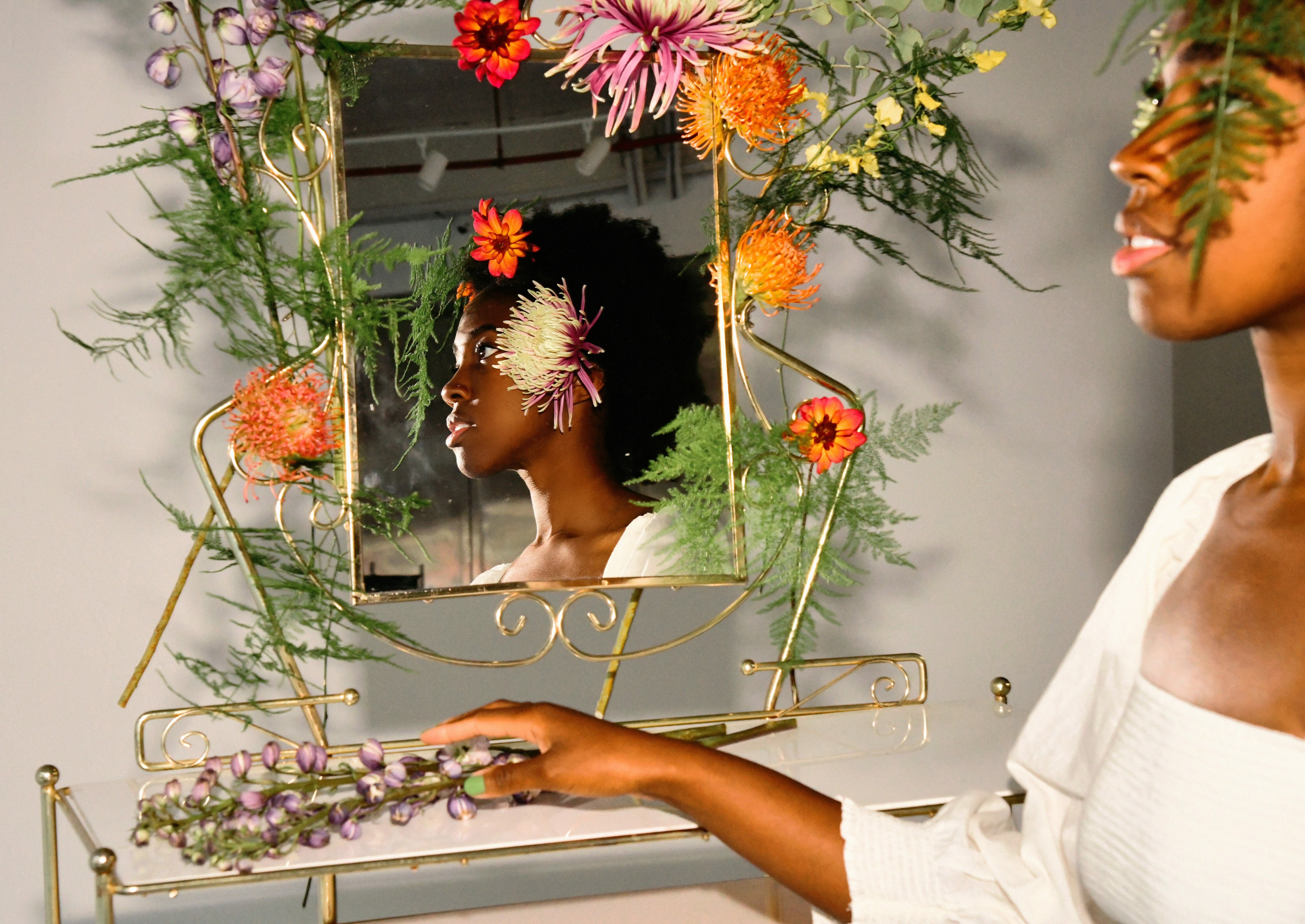 Reflection of a person with flowers, leaves, and foliage surrounding their face in a mirror.