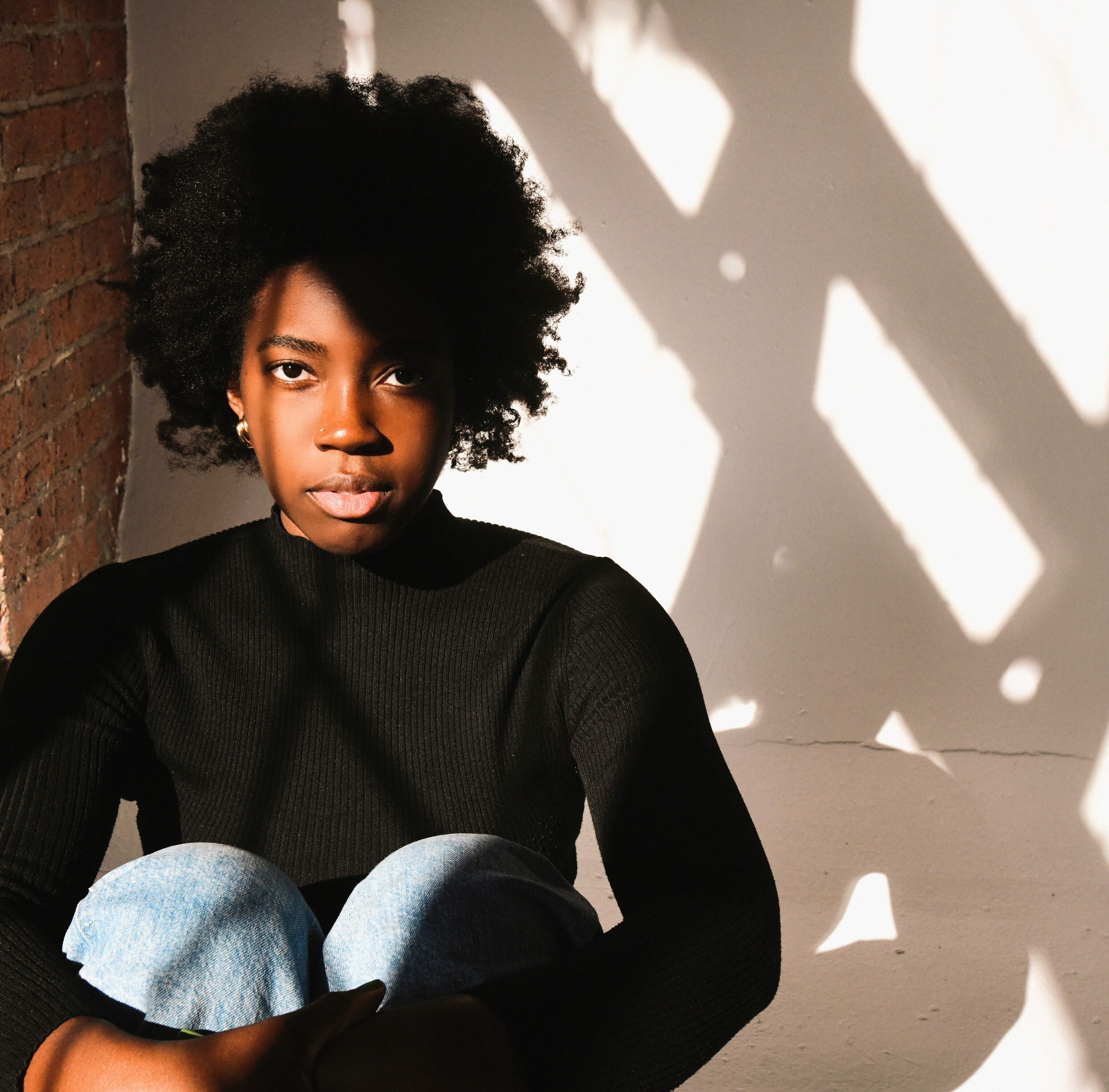 Image shows a Black person with curly hair wearing a black top, sitting against a brick wall with sunlight and shadow patterns on the wall behind them.