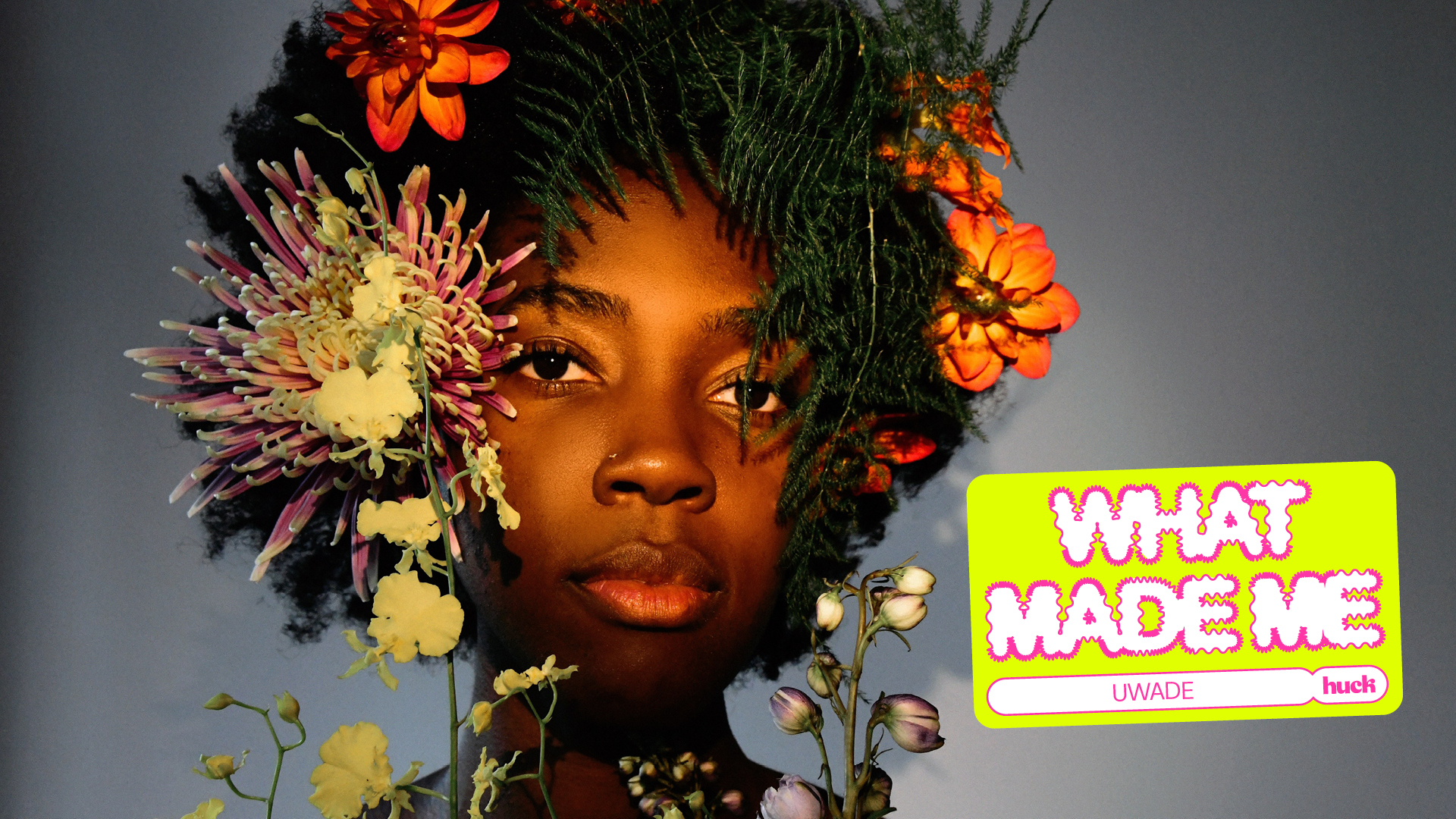 A person's face surrounded by colourful flowers. The flowers include orange, red, and yellow dahlias, as well as smaller yellow blooms. The person's expression is serious.