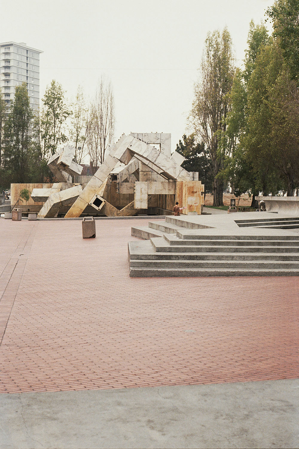 Angular metal sculpture with bronze and grey geometric forms on brick plaza, surrounded by trees and modern buildings.