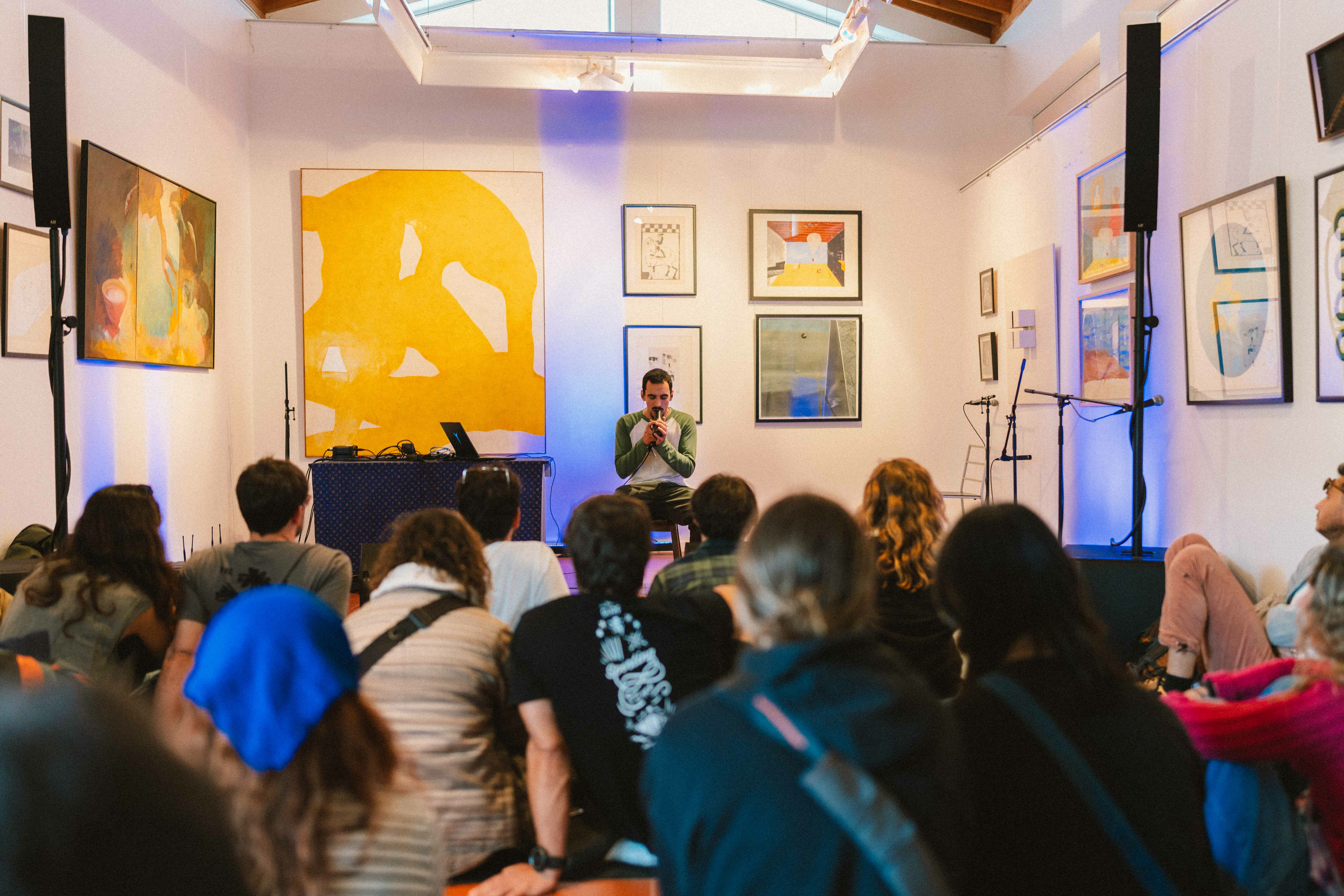 A crowded indoor event with a speaker on stage and a large yellow artwork on the wall behind them.