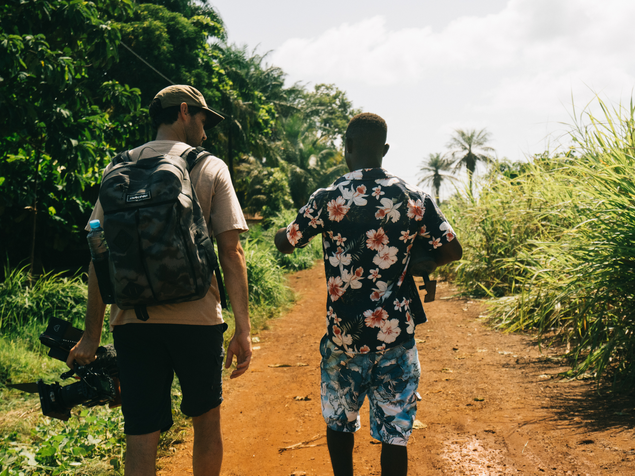Two people walking away on dirt path through lush tropical vegetation, one wearing backpack and cap, other in floral shirt.
