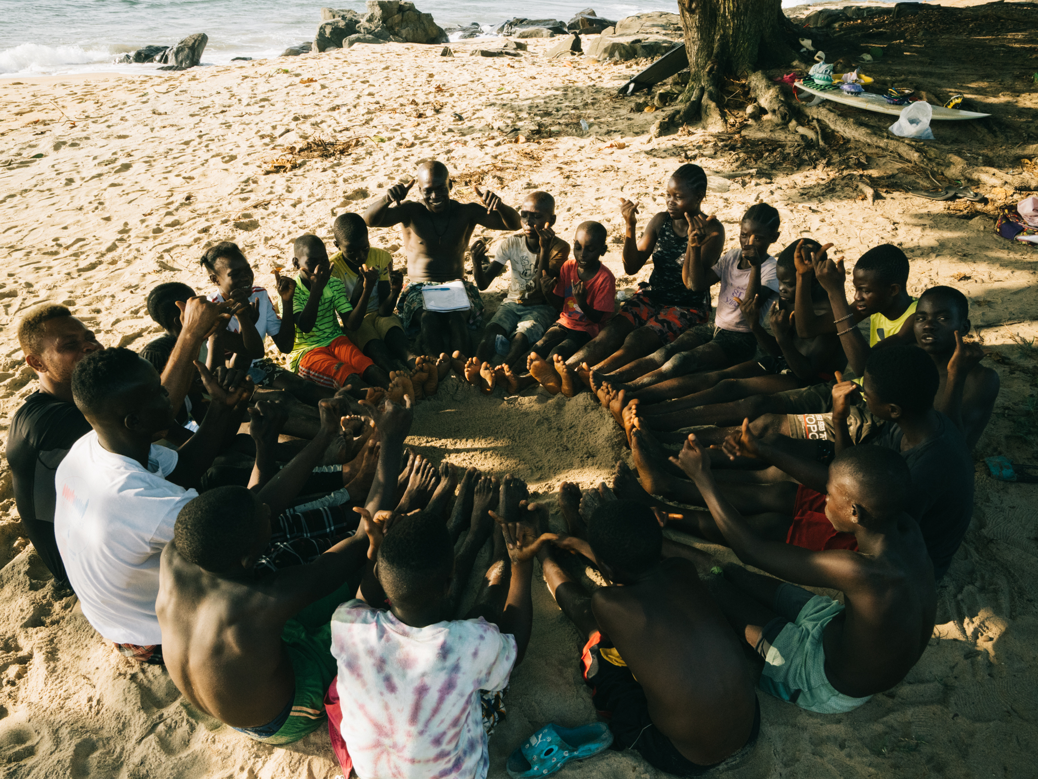 Large group of children and adults sitting in circle on sandy beach under palm tree shade, with ocean and rocks visible in background.