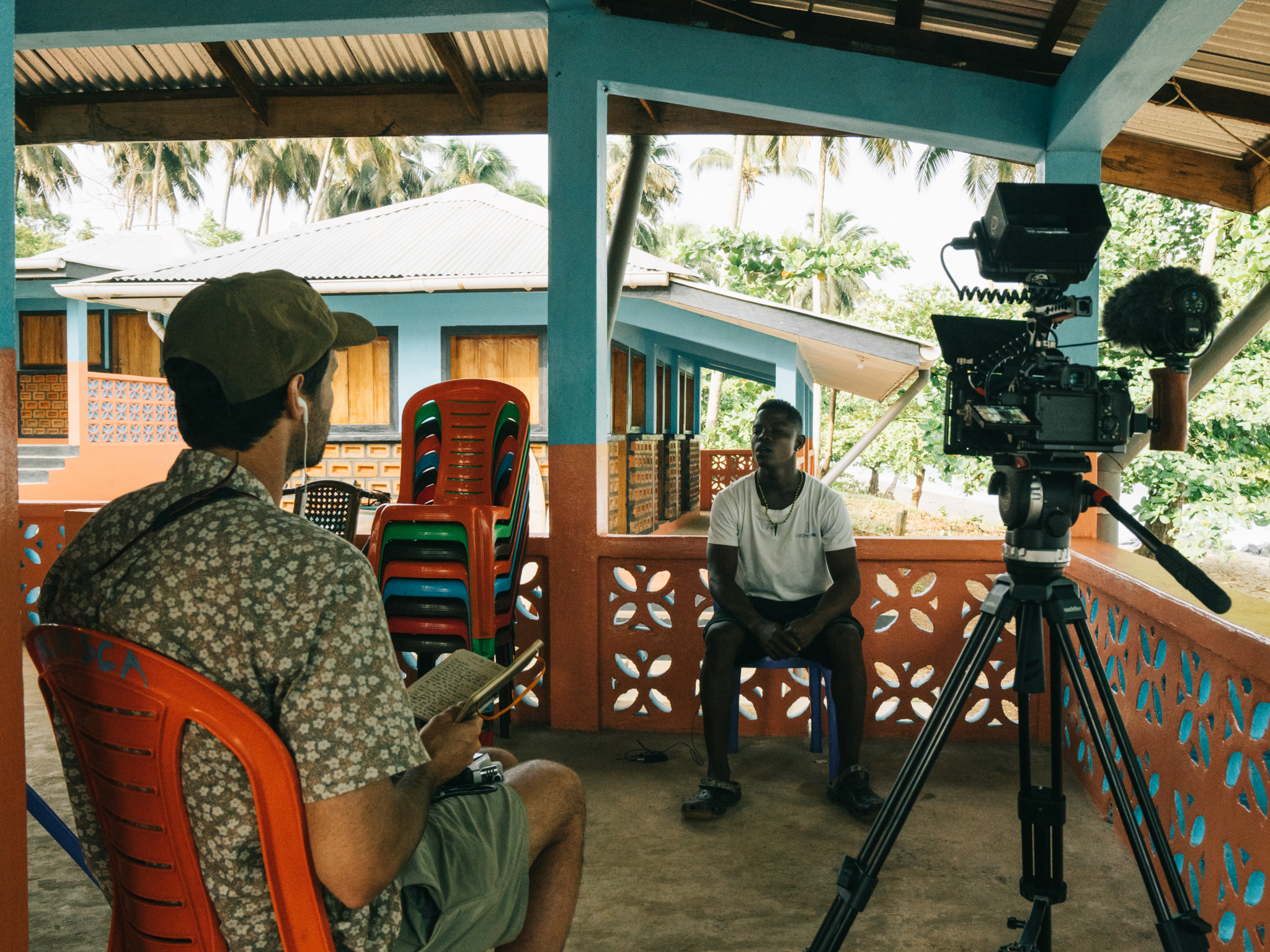 Two men on covered porch with professional camera on tripod. Red plastic chairs, decorative railing, tropical buildings and palm trees visible in background.