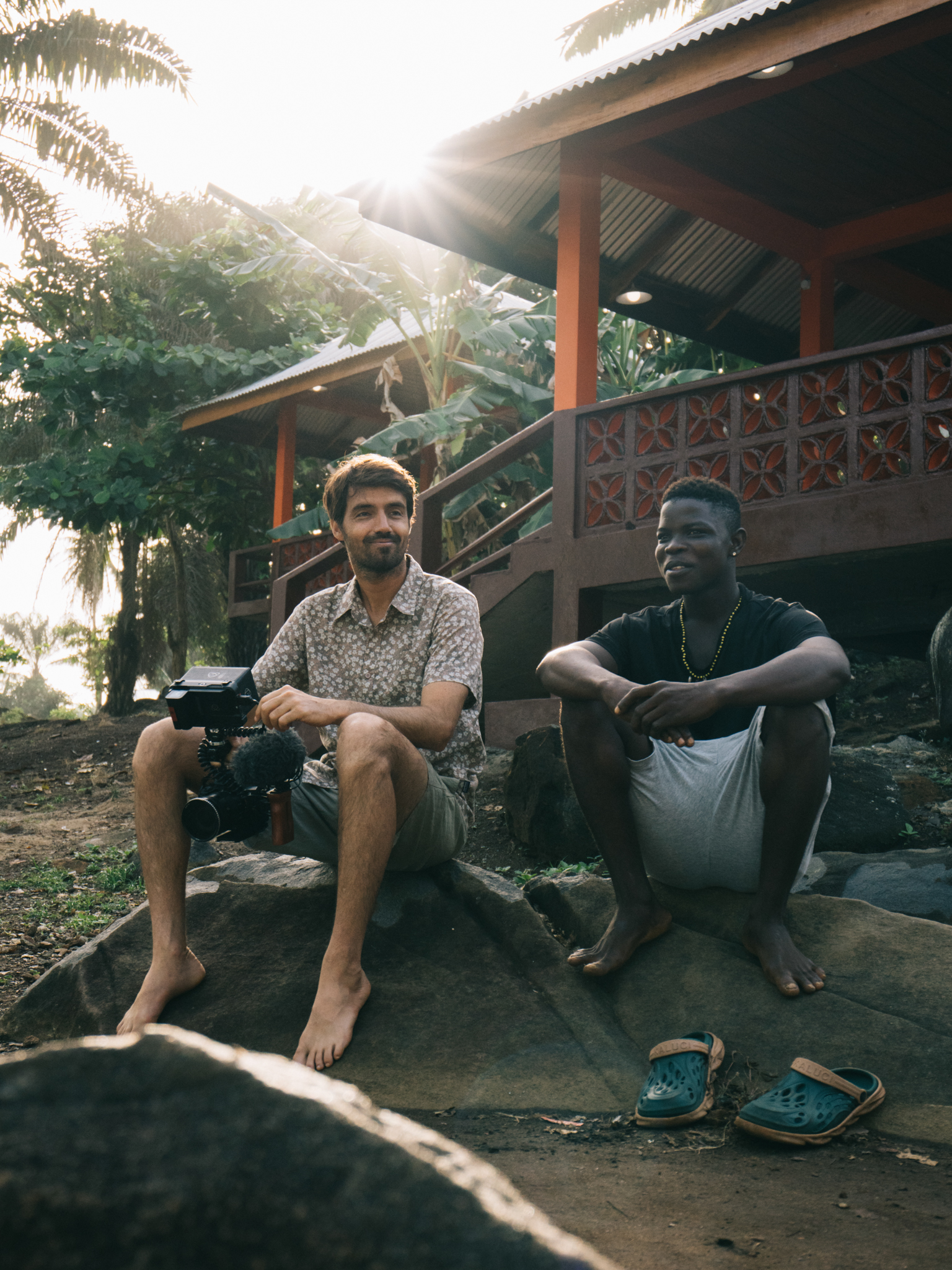 Two men sitting on concrete steps outside wooden building with corrugated roof, tropical plants and bright sunlight in background.