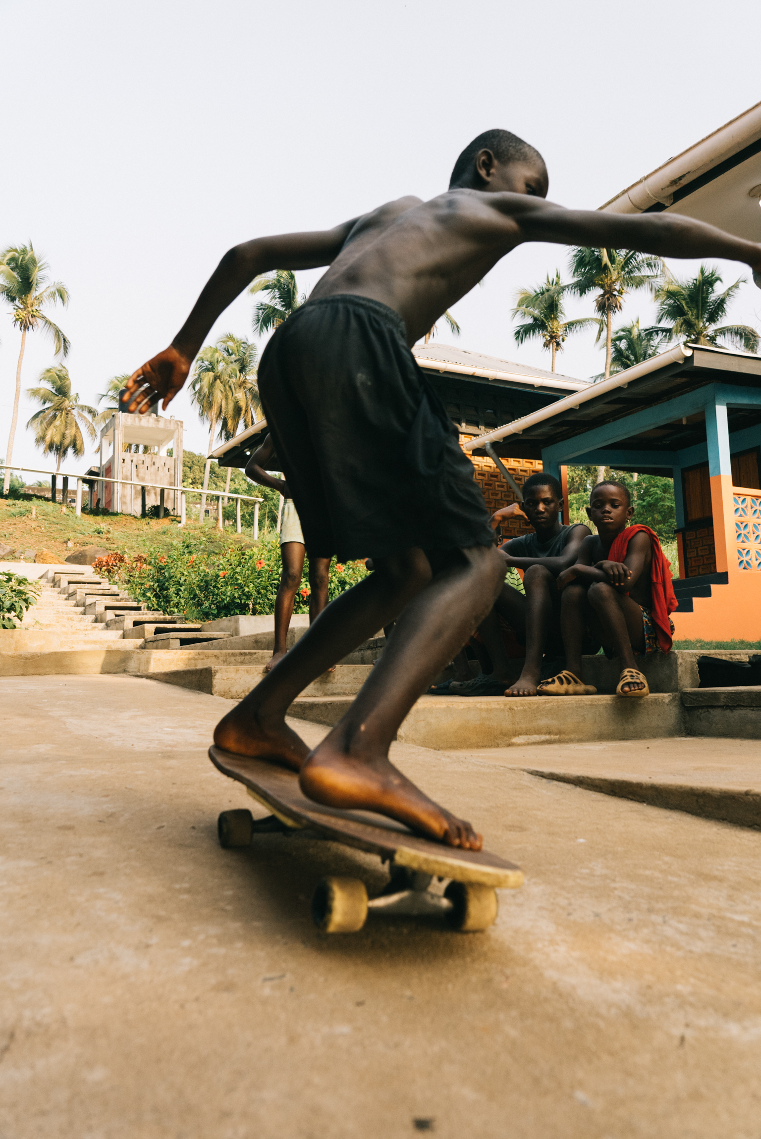 Young man skateboarding on concrete steps with palm trees and colourful buildings in background, other people sitting nearby.