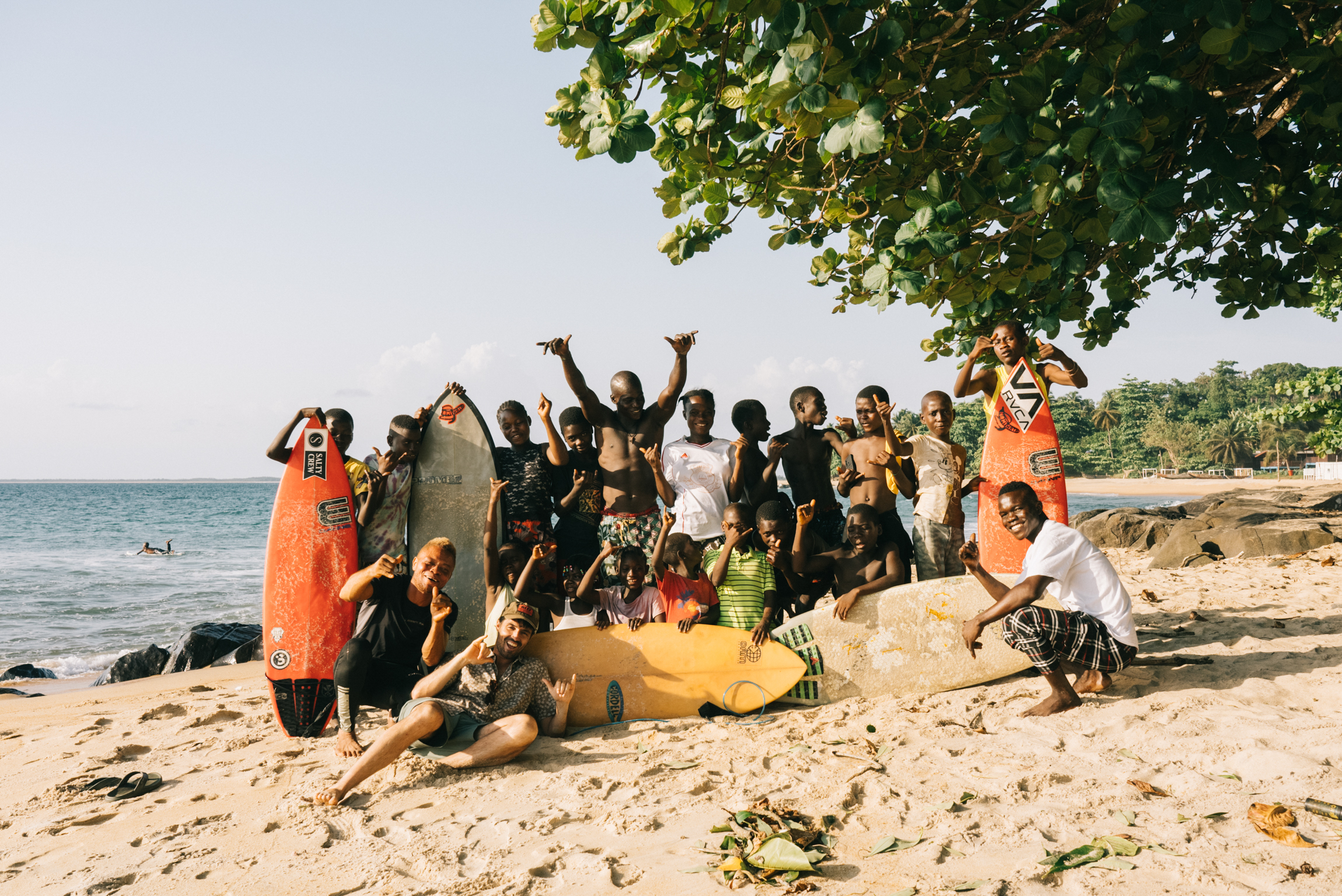 Large group of people posing on sandy beach with surfboards, some with raised arms, green tree overhead, ocean and coastline visible.
