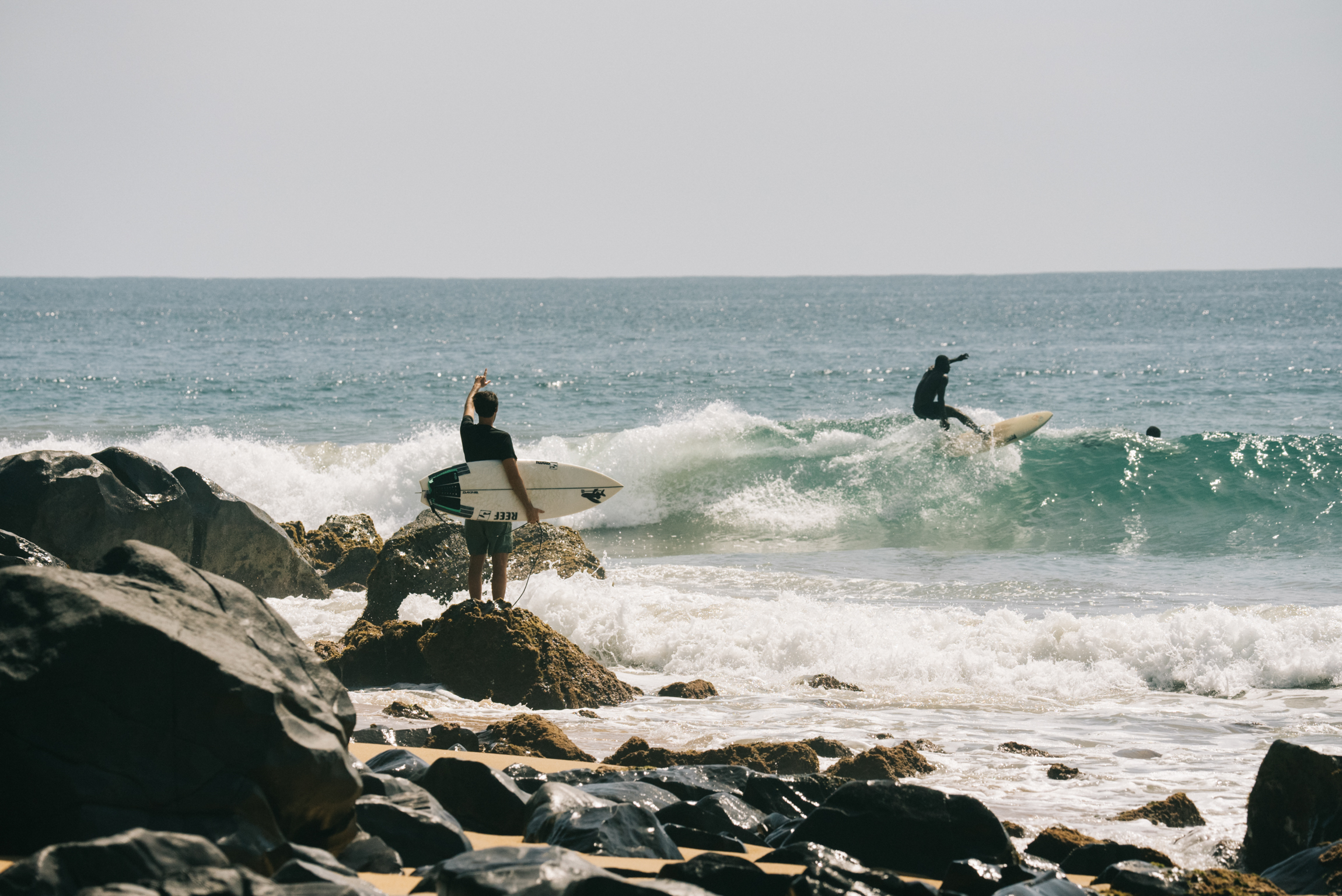 Two people with surfboards on rocky coastline, one standing on rocks, another in white-capped waves under overcast sky.