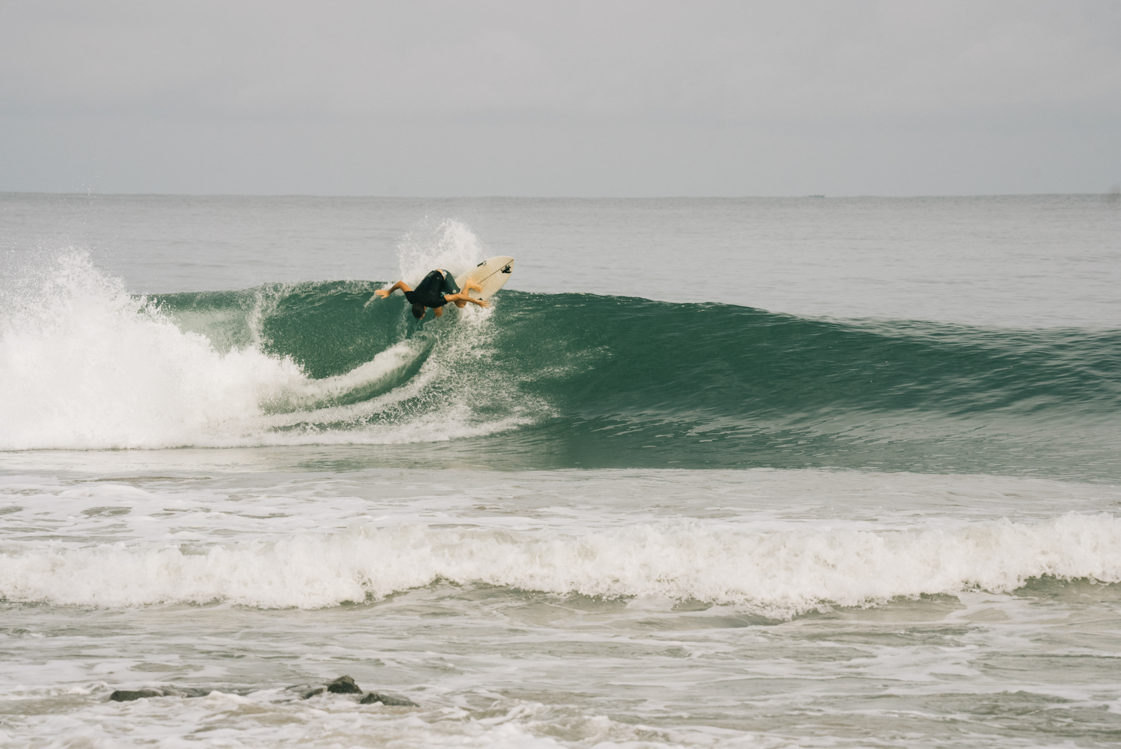 Surfer riding green wave with white foam spray against grey cloudy sky and choppy ocean water.