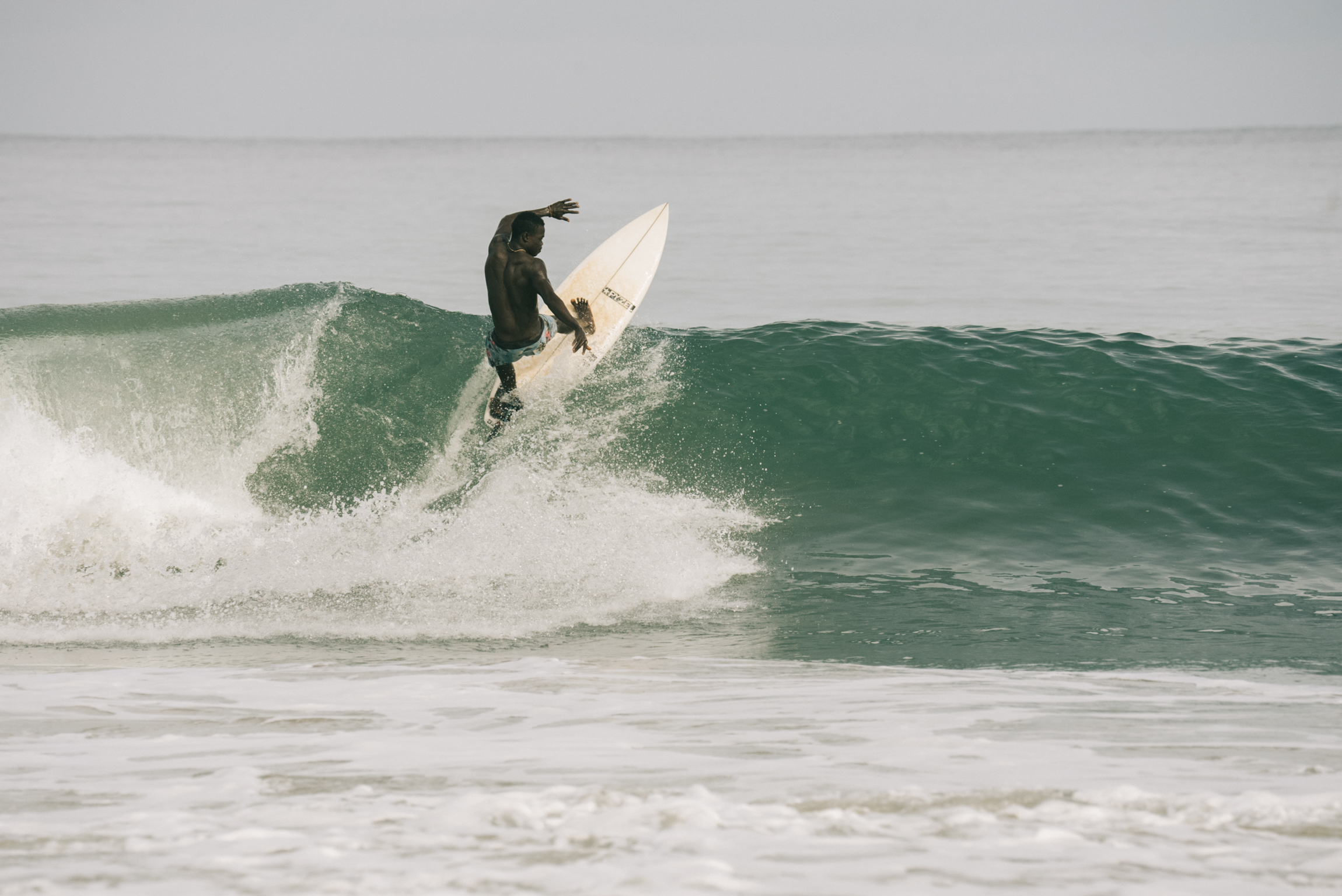 Surfer in dark wetsuit riding green wave with white foam, holding white surfboard vertically whilst performing manoeuvre.