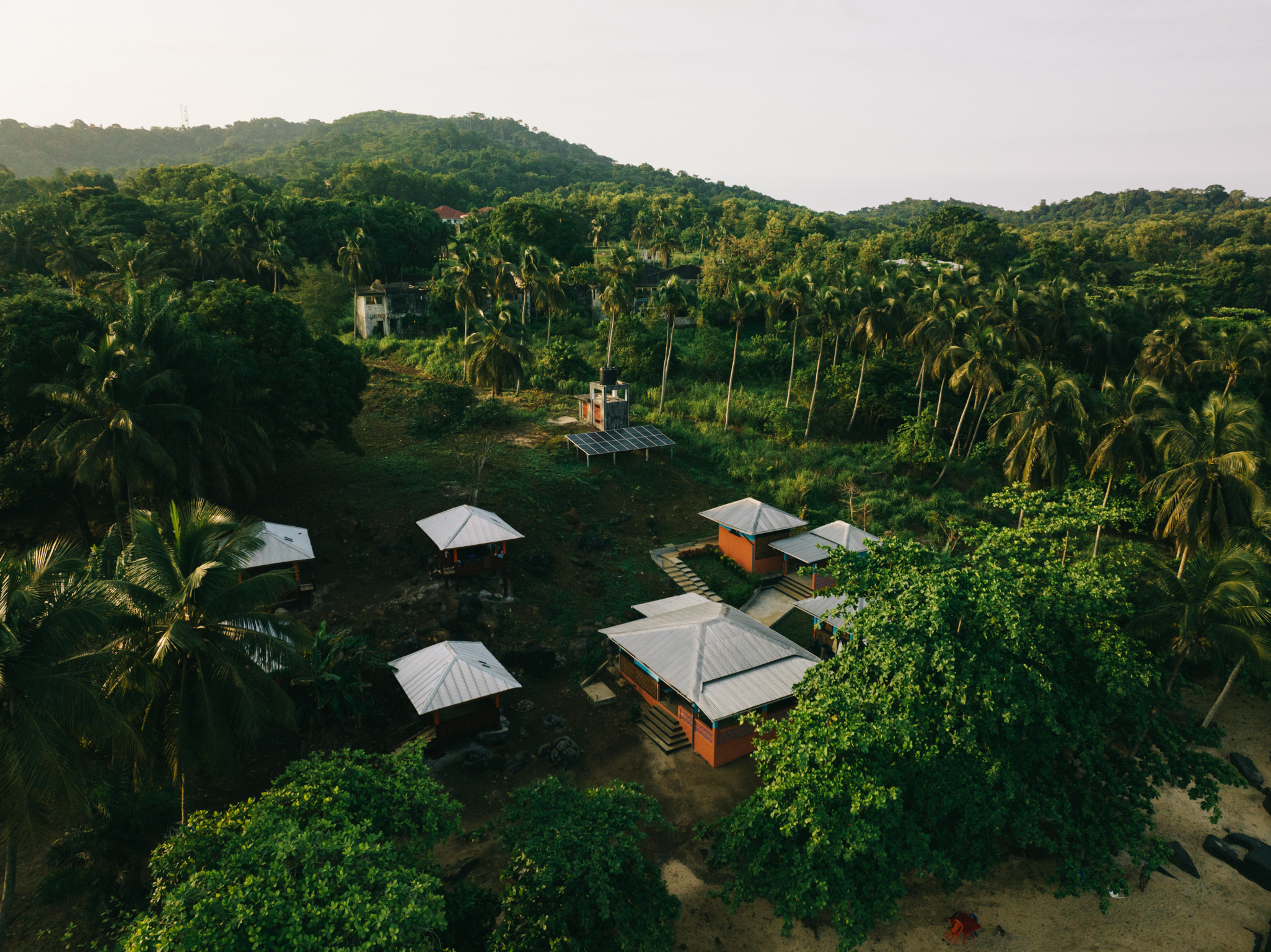 Aerial view of small houses with metal roofs scattered amongst dense green tropical vegetation and forested hills.