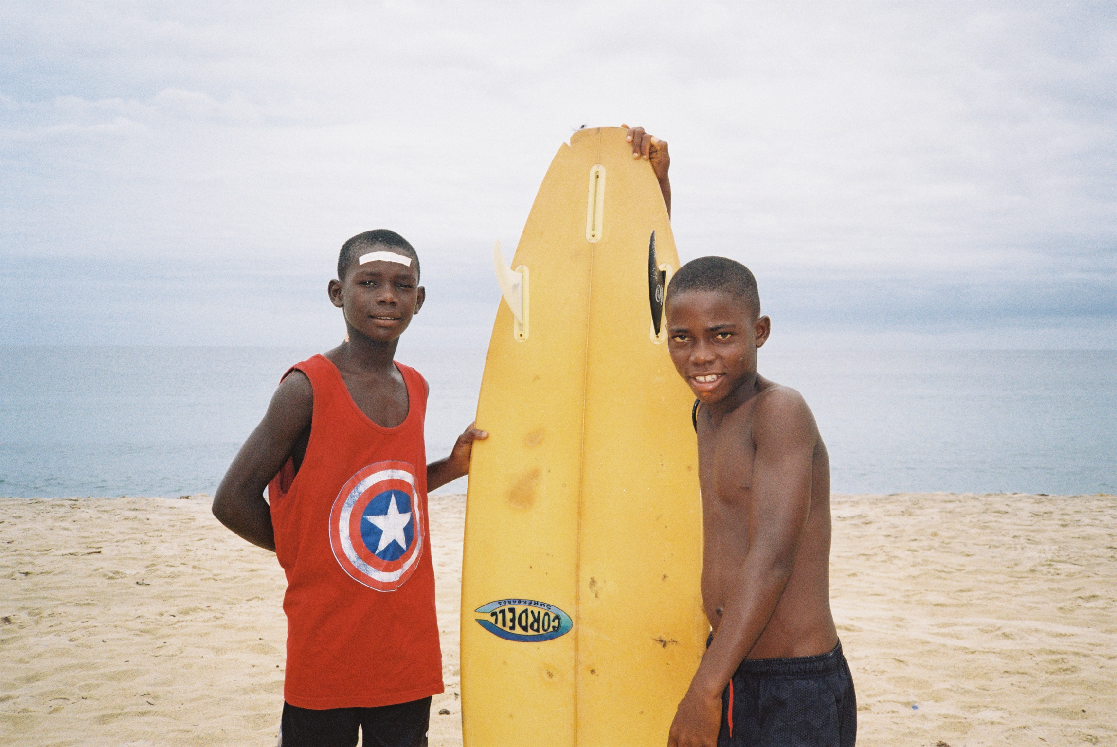 Two boys on beach with yellow surfboard. Left boy wears red Captain America vest, right boy shirtless. Cloudy sky and ocean in background.