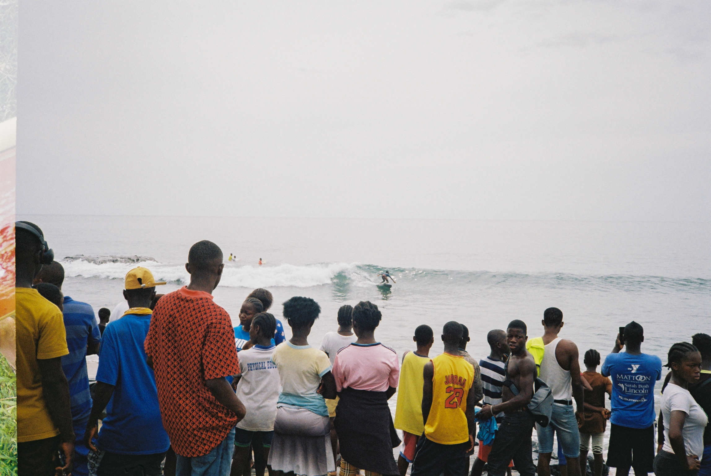 Group of people in colourful clothing standing on beach watching waves and helicopter in distance under overcast sky.