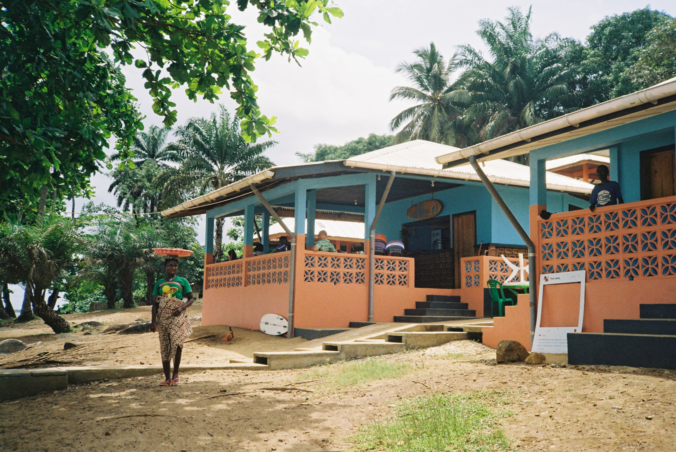 Orange and turquoise beach bungalows with decorative block walls beneath palm trees on sandy ground.