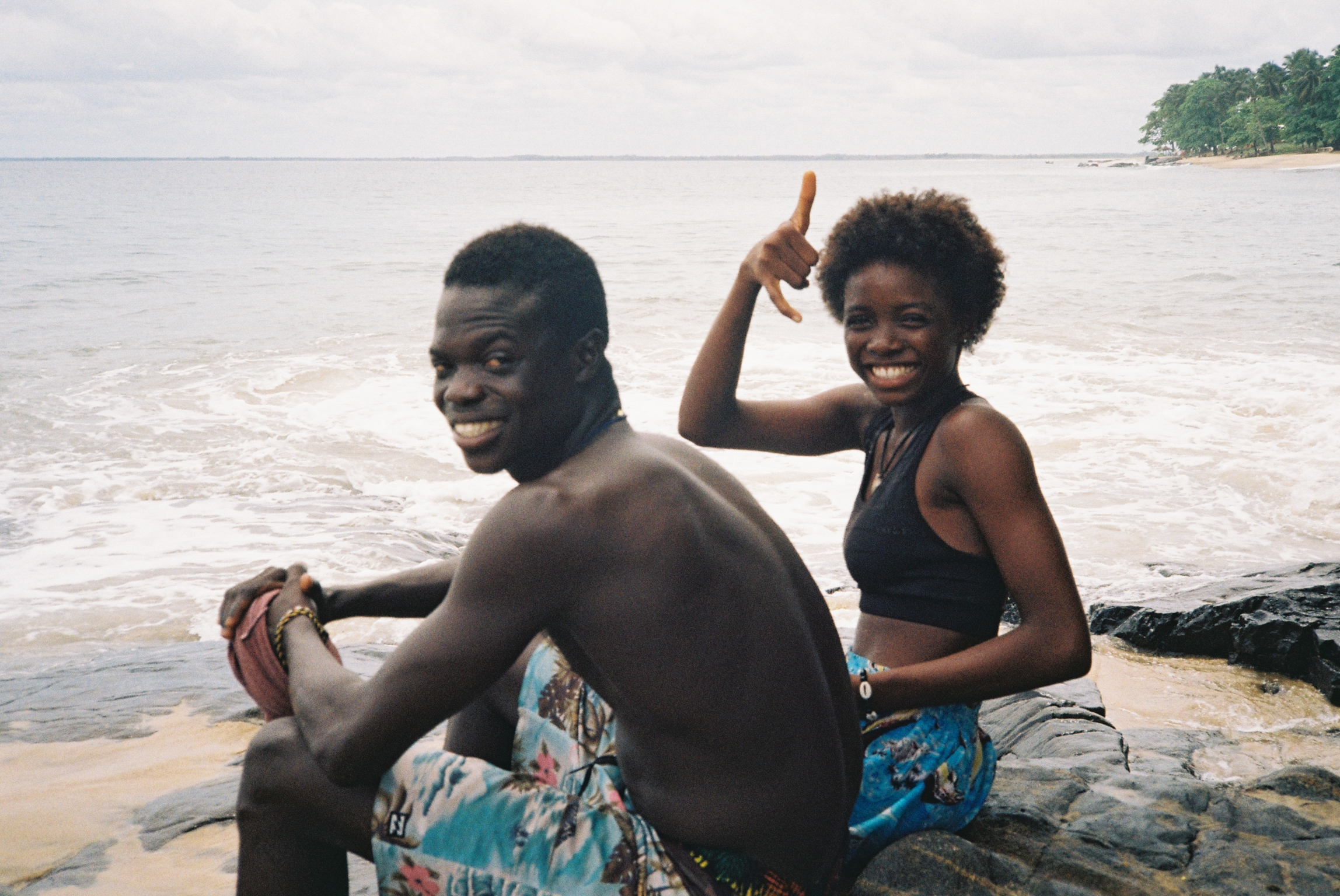 Two people sitting on rocks by the sea, one shirtless man and one woman in swimwear giving thumbs up, with waves and shoreline behind them.