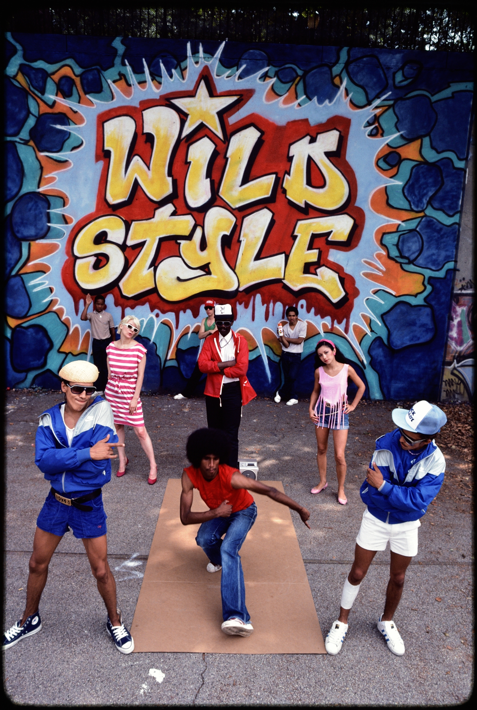 Group of young people breakdancing in front of graffiti mural reading "WILD STYLE" in yellow and red letters with blue decorative elements.