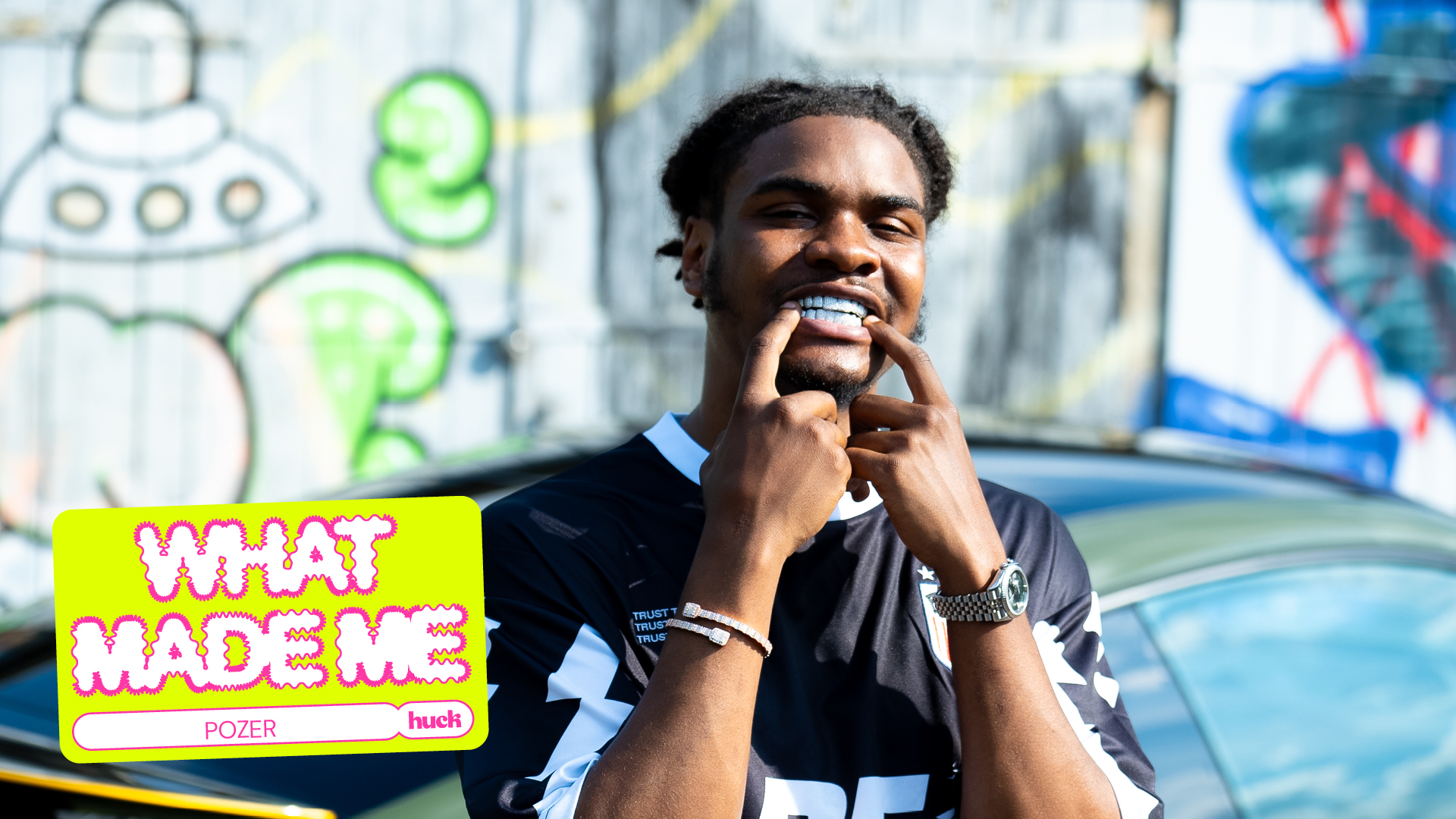 Black man in striped shirt holding microphone, smiling. Colourful graffiti wall behind. Yellow "WHAT MADE ME" text overlay bottom left.