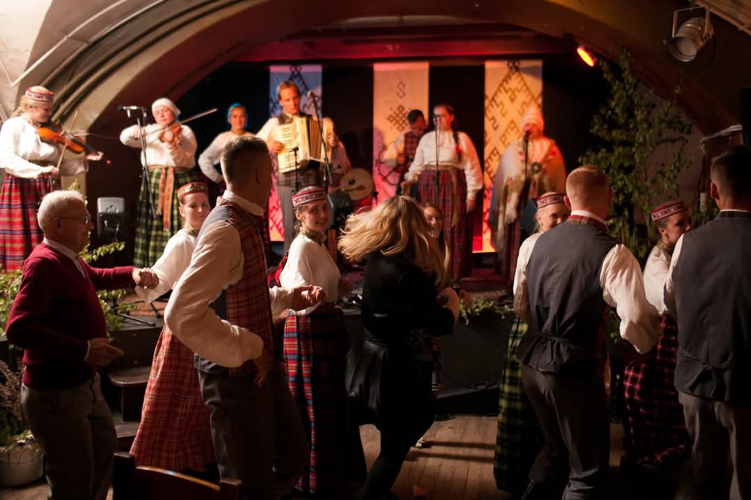 Lively crowd of people wearing traditional Scottish attire, including kilts, dancing and playing music on a stage.