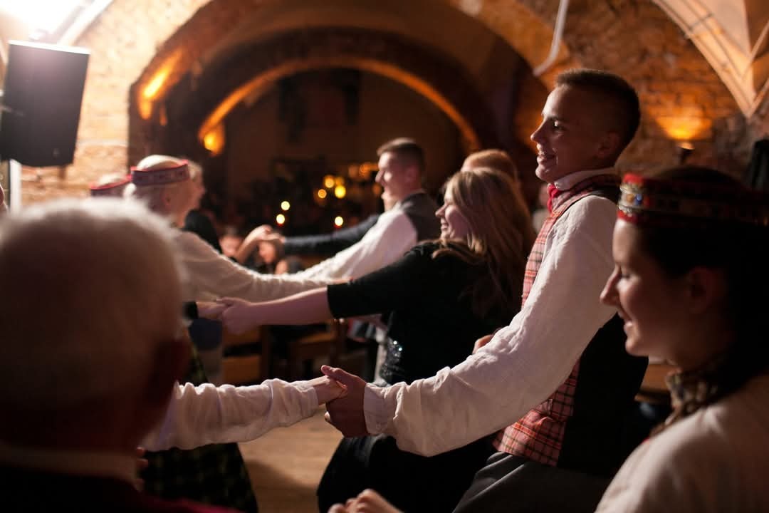 A group of people dancing at a social event in a dimly lit room with an arched stone doorway visible in the background.