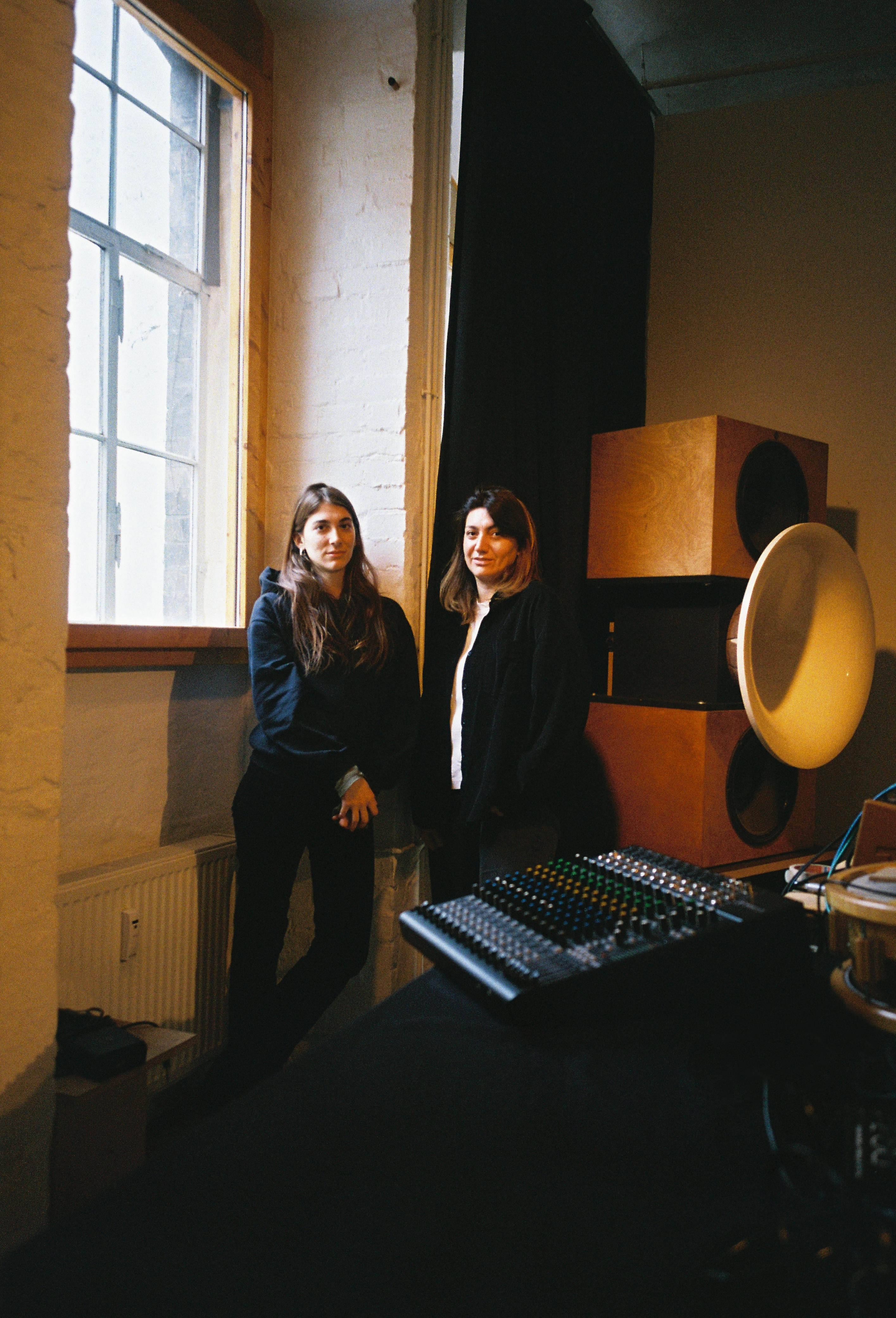 Two women in dark clothing standing by large window in recording studio with mixing desk and monitors visible.