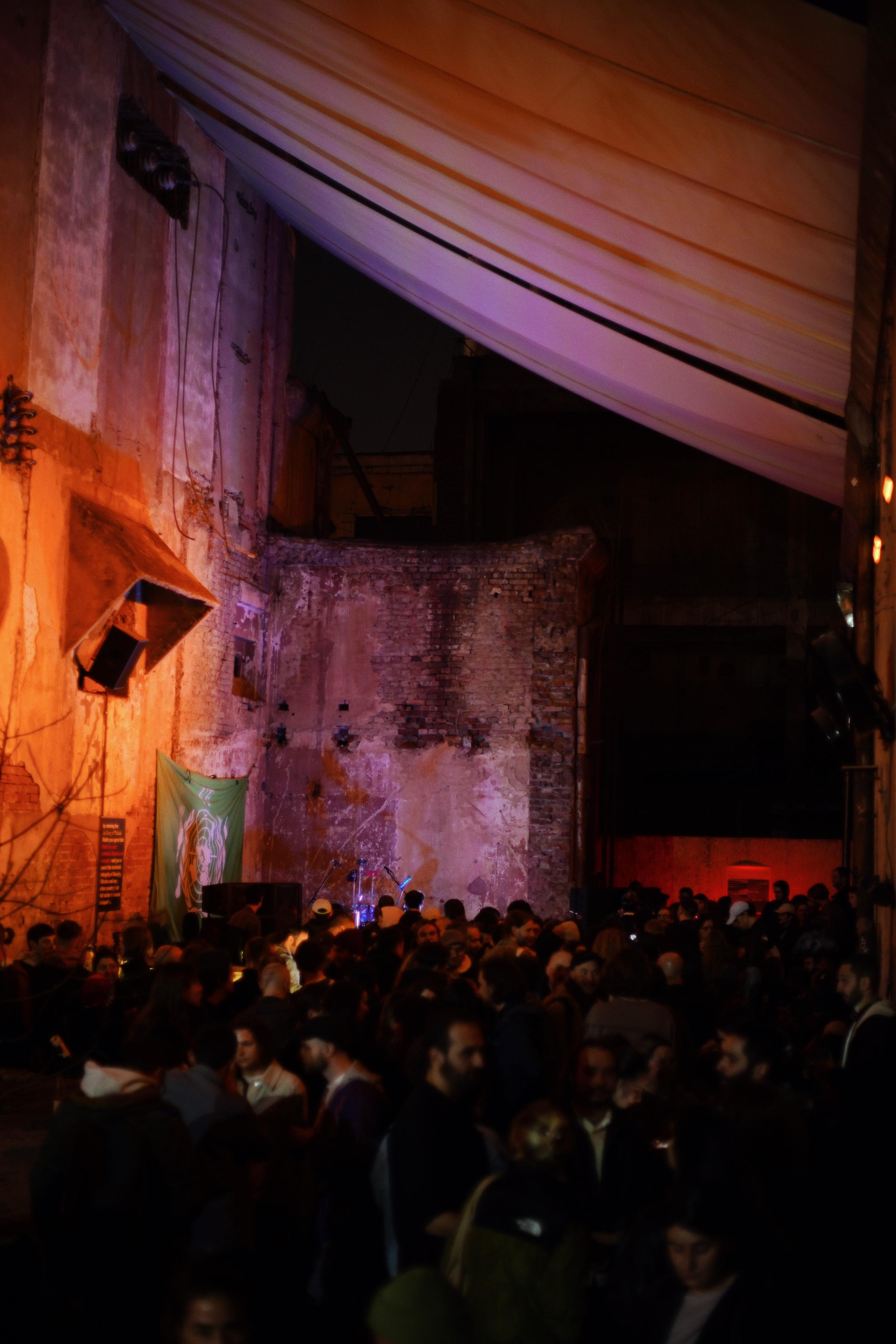 Concert venue interior with crowd of people in foreground, brick walls lit with purple and orange lighting, exposed ceiling beams above.