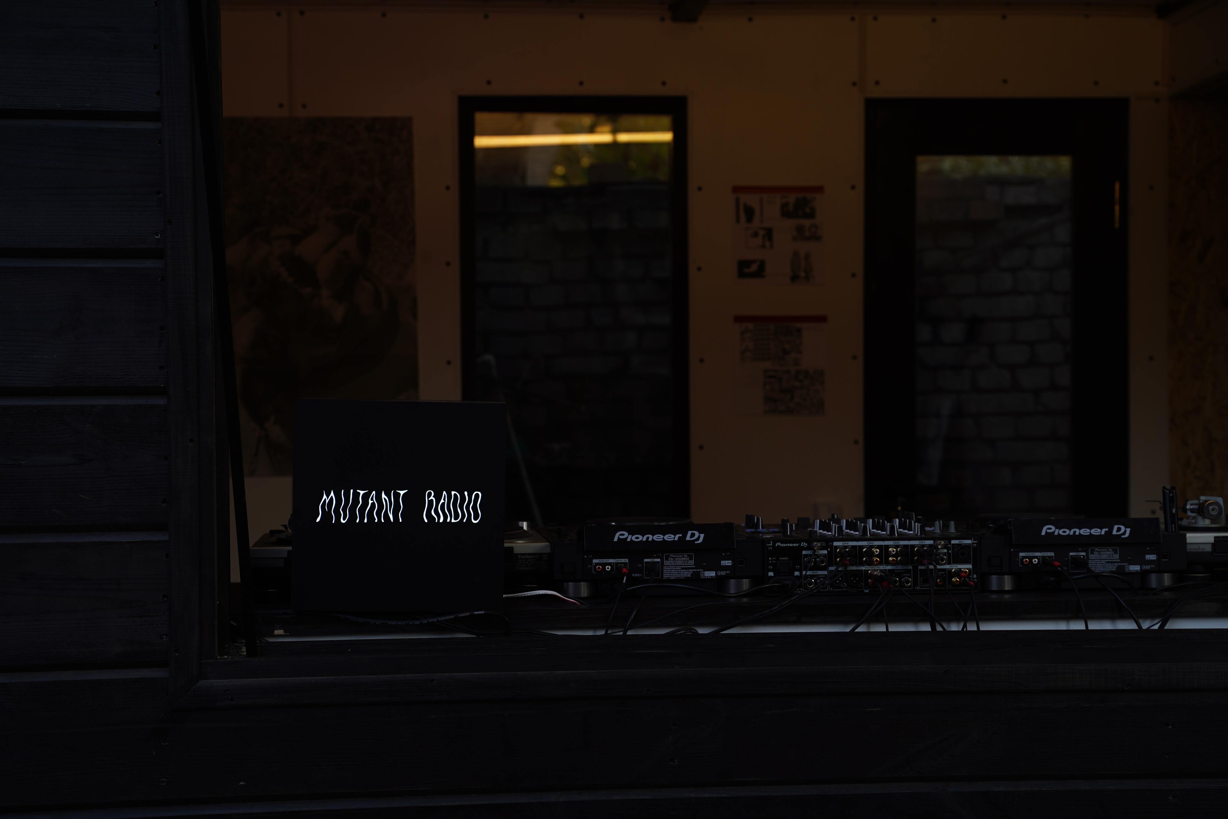 Dark storefront at night with "MUTANT RADIO" sign in white letters, two doorways with warm light above, bicycles parked outside.