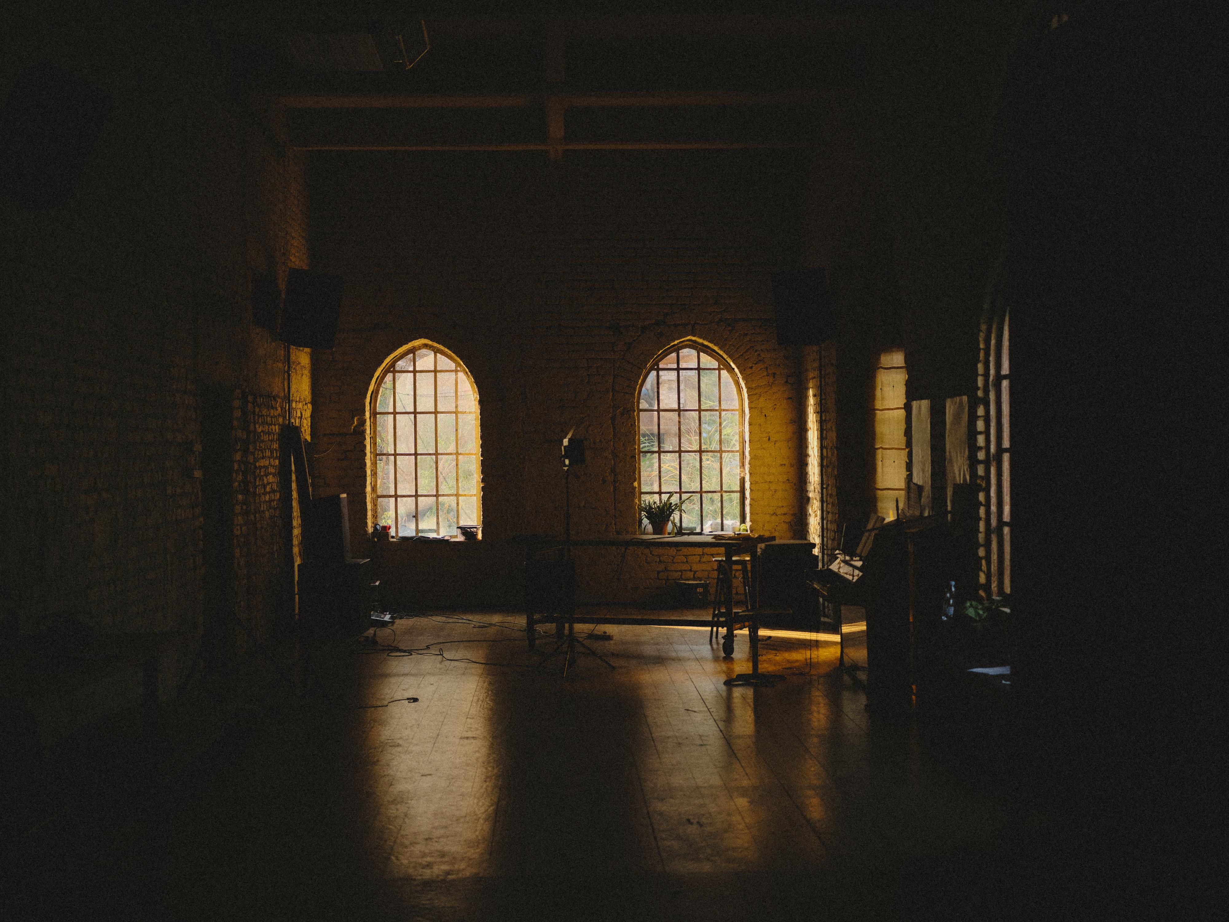 Dimly lit interior with two arched windows casting golden light onto wooden floor, stone walls, wooden furniture visible in shadows.