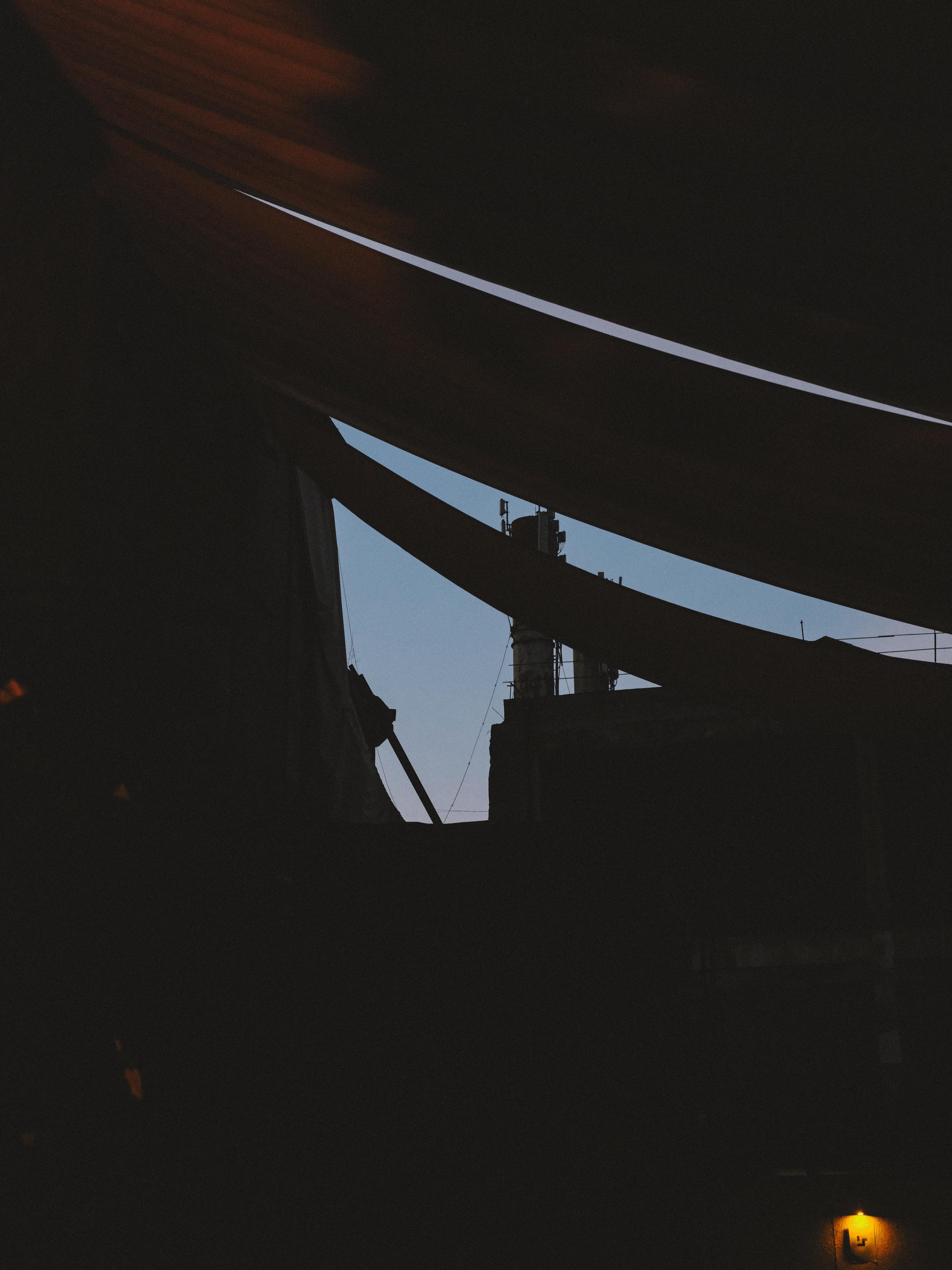 Dark interior space with curved concrete or metal beams creating angular shadows. Light blue sky visible through geometric opening. Small yellow light source in bottom corner.